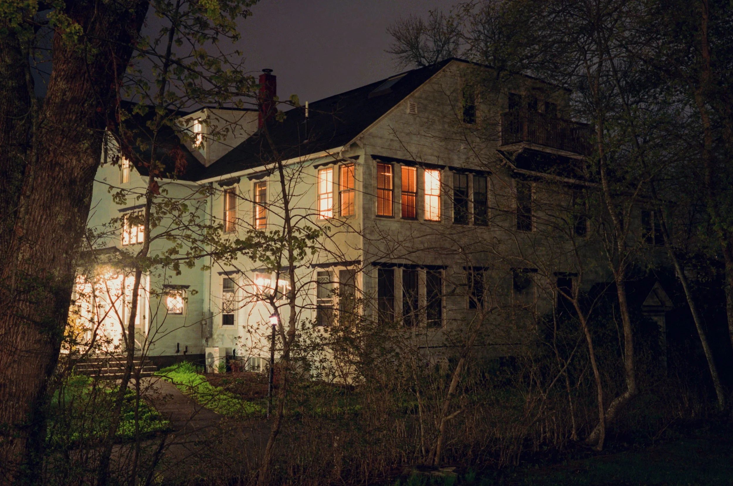 A multi-story house at night with warm light glowing from its windows, surrounded by leafless trees and a dark sky.