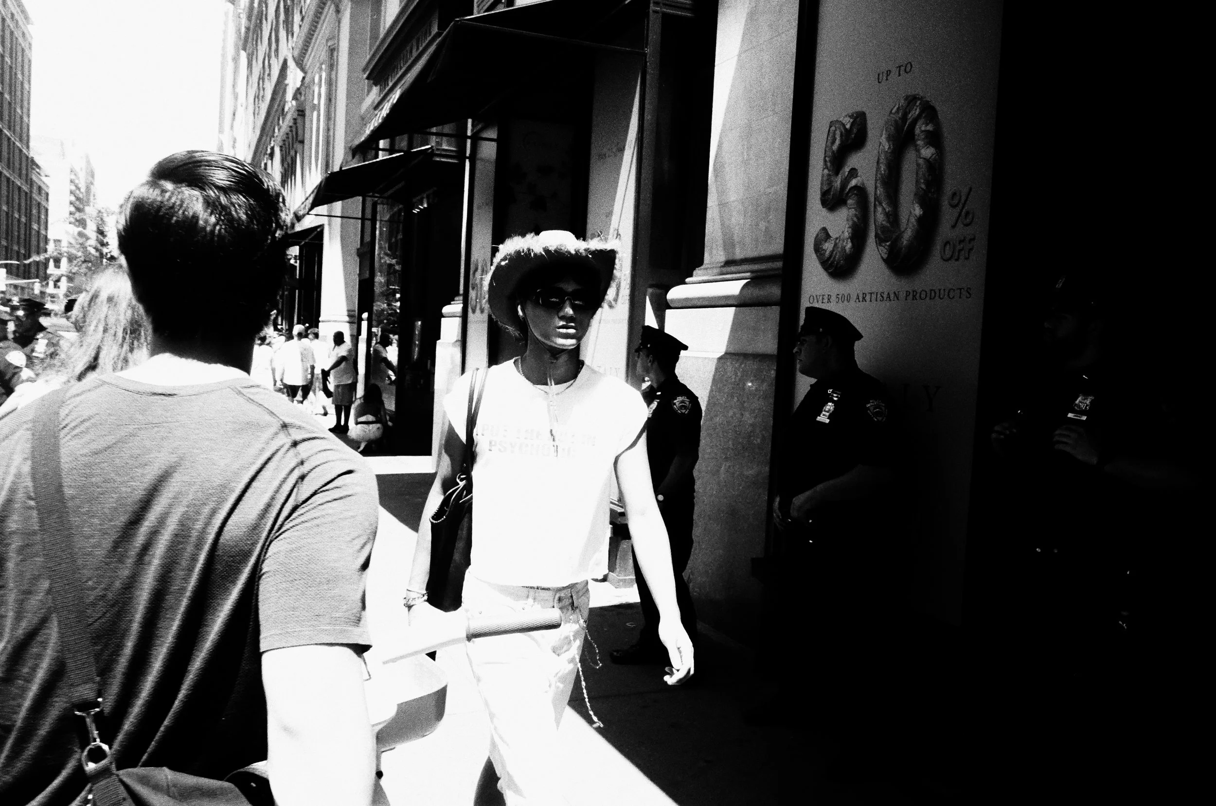 A street scene in black and white featuring two women in conversation. One woman, with her back to the camera, has her hair tied back and is wearing a long-sleeved shirt. The other woman, facing the camera, is wearing sunglasses, a wide-brimmed hat, 