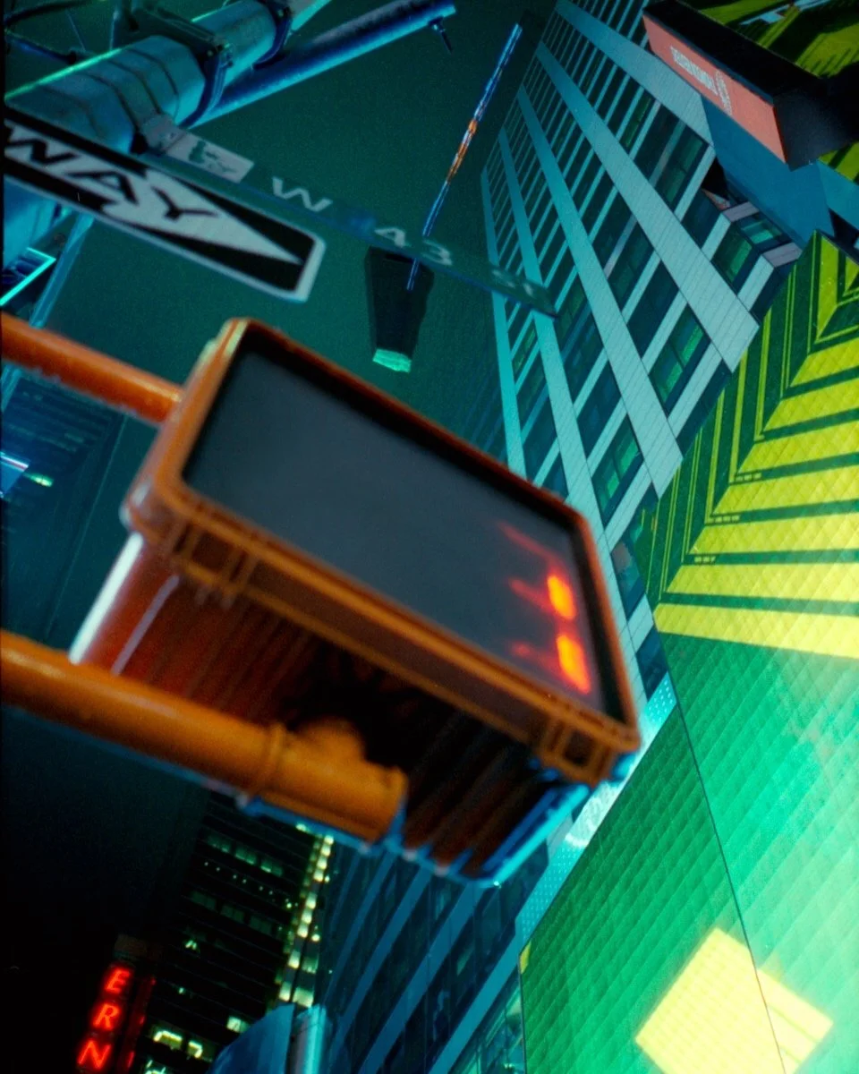Looking up at a tall city building at night, with bright, colorful digital billboards and street signs visible. A street sign reading 'WAY' can be seen, along with illuminated building windows.