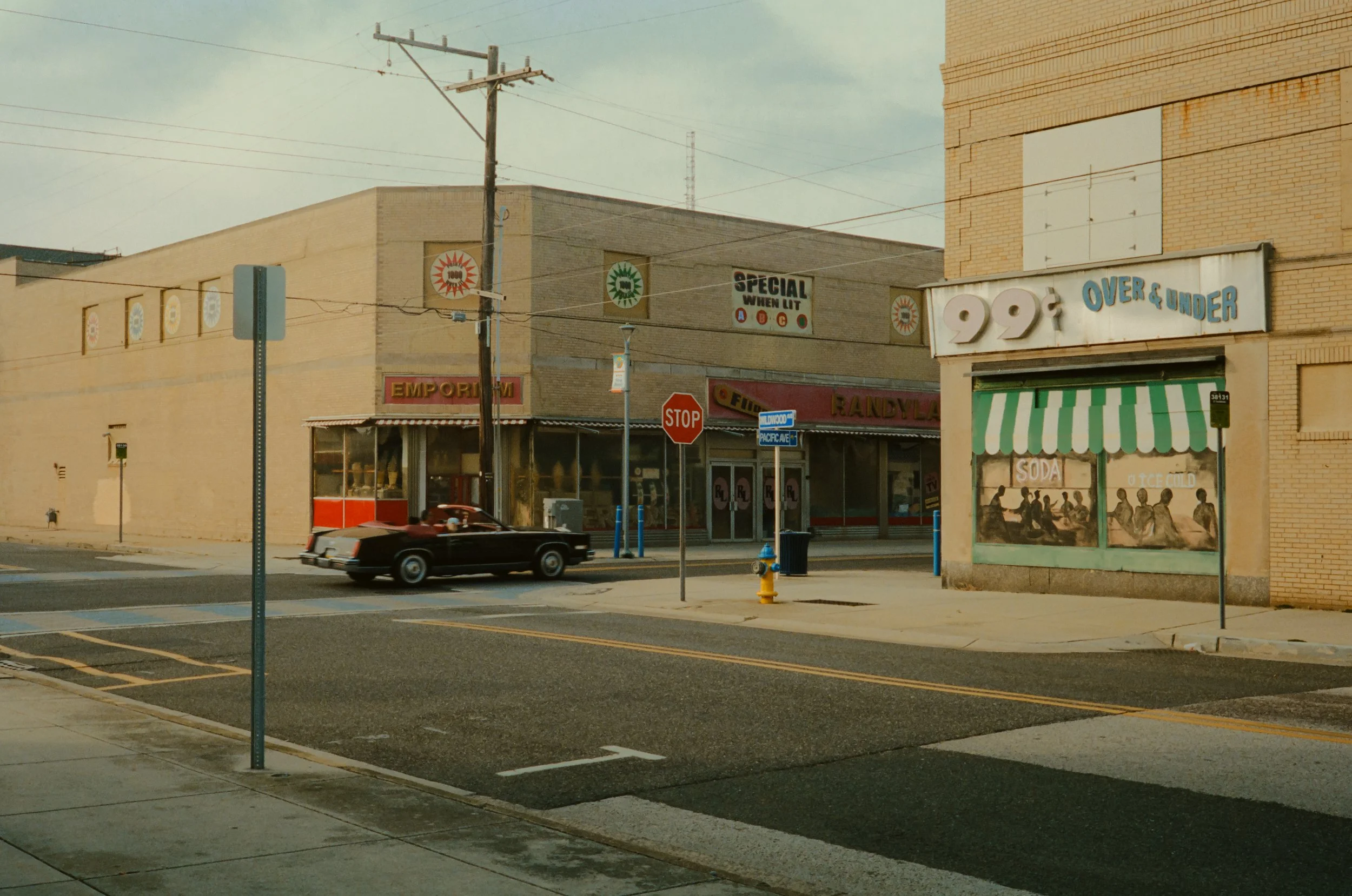 A city street scene with a black car driving past a building with signs advertising a dollar store, special offers, and an emporium. Utility poles and power lines are visible, and the sky is overcast.