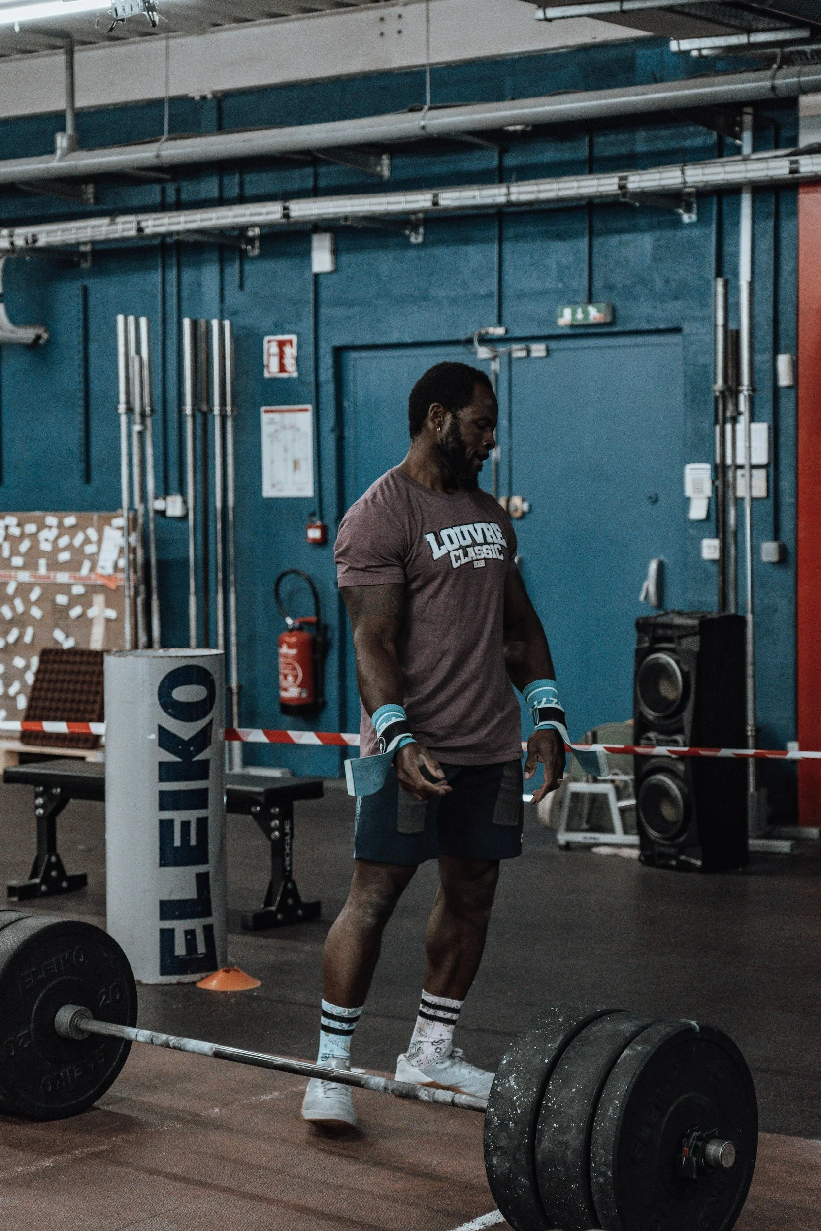 A manreparing for a deadlift, holding lifting straps in each hand. In front of him on the floor is a loaded barbell and weight plates. The gym has blue walls, and there is gym equipment such as a pull-up bar and a fire extinguisher in the background.