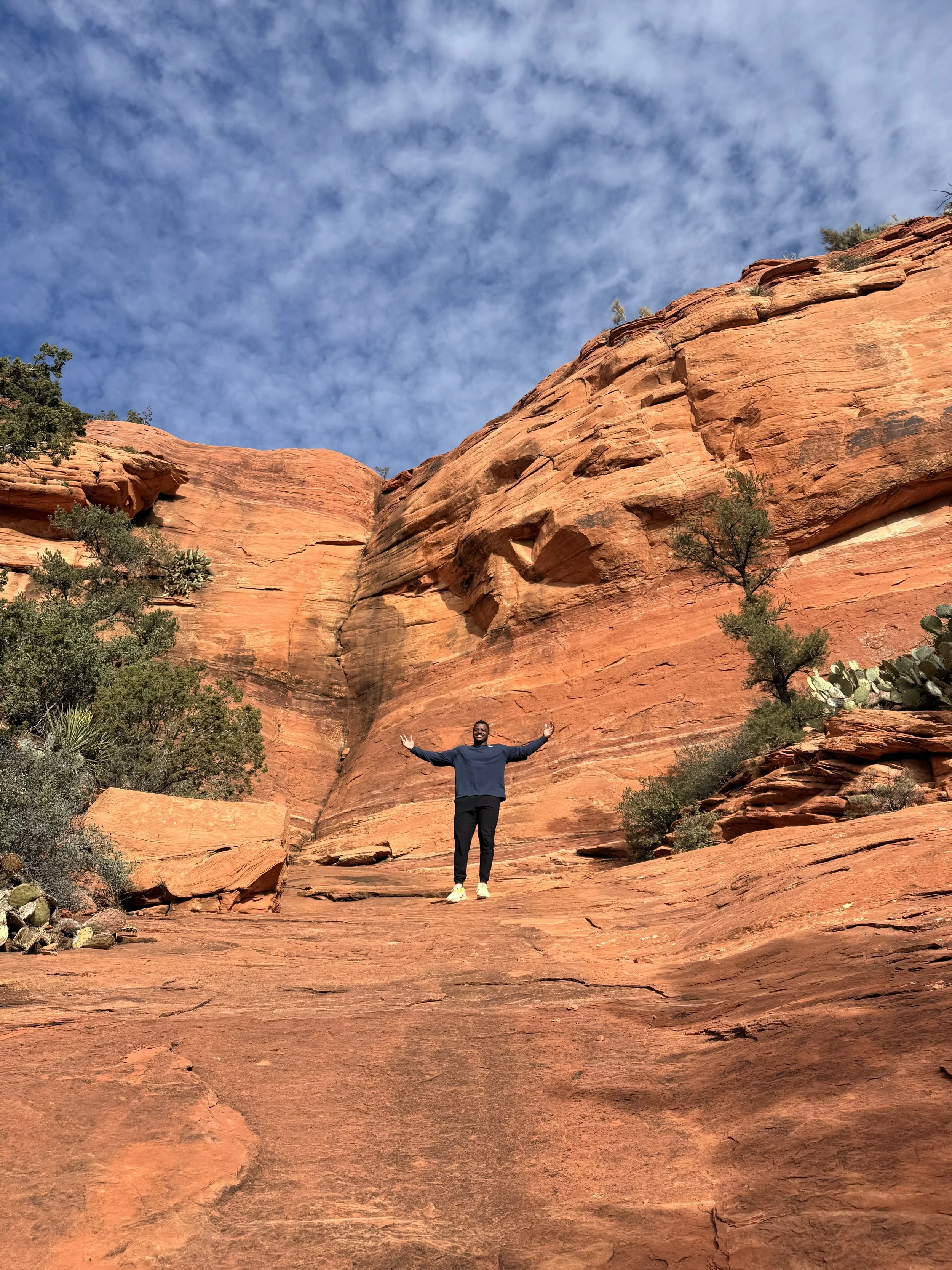 Andre Whitely standing with arms outstretched in front of red rock formations and blue sky with clouds.