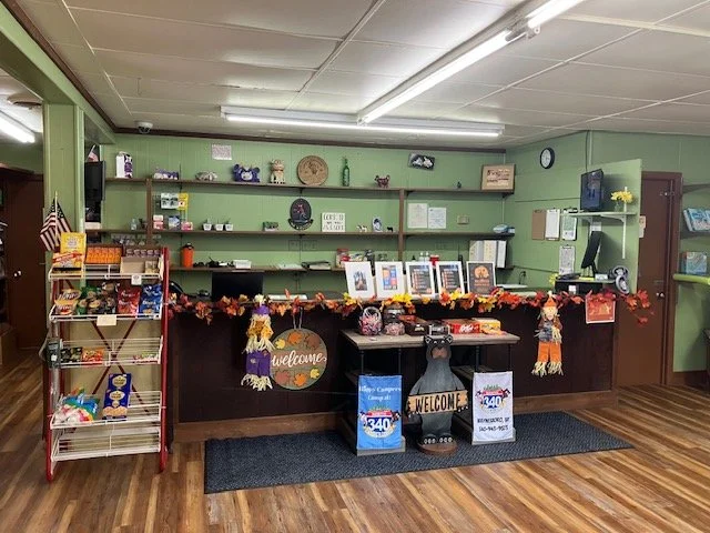 Indoor reception area with a wooden counter decorated with autumn leaves, pumpkins, and welcome signs, against light green walls with shelves containing decorative items, books, and a TV.