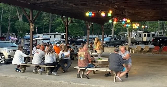 A group under pavilion with string lights overhead and picnic tables with RVs in the background.