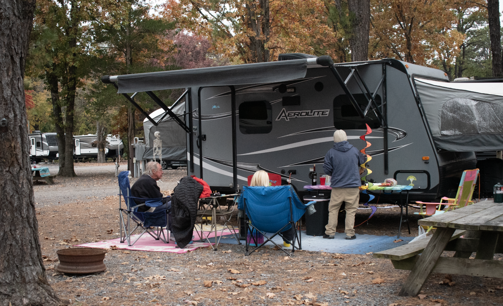 A family sitting in chairs around their RV site with a Fire ring and fall trees and foliage.