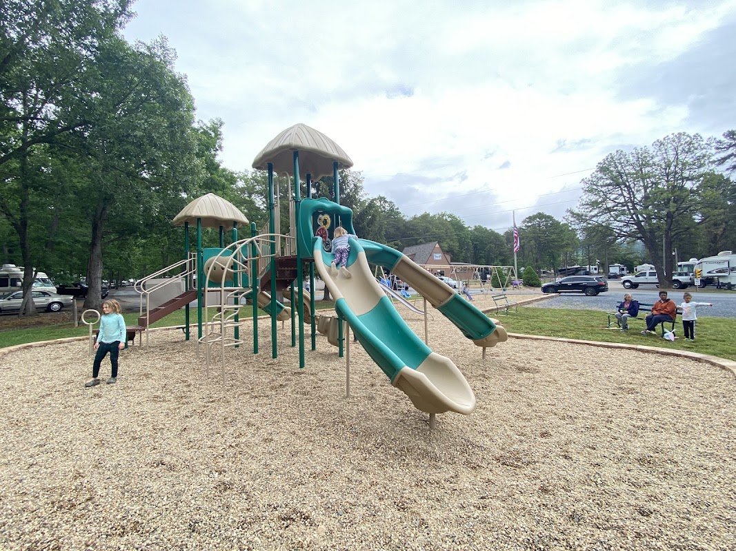 Children playing on a playground with slides, climbing structures, and shaded towers, surrounded by trees and a parking lot.
