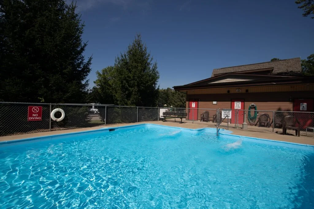 An outdoor swimming pool with blue water, surrounded by a chain-link fence with a no diving sign, next to a building with chairs and equipment, set against a background of green trees and a clear blue sky.