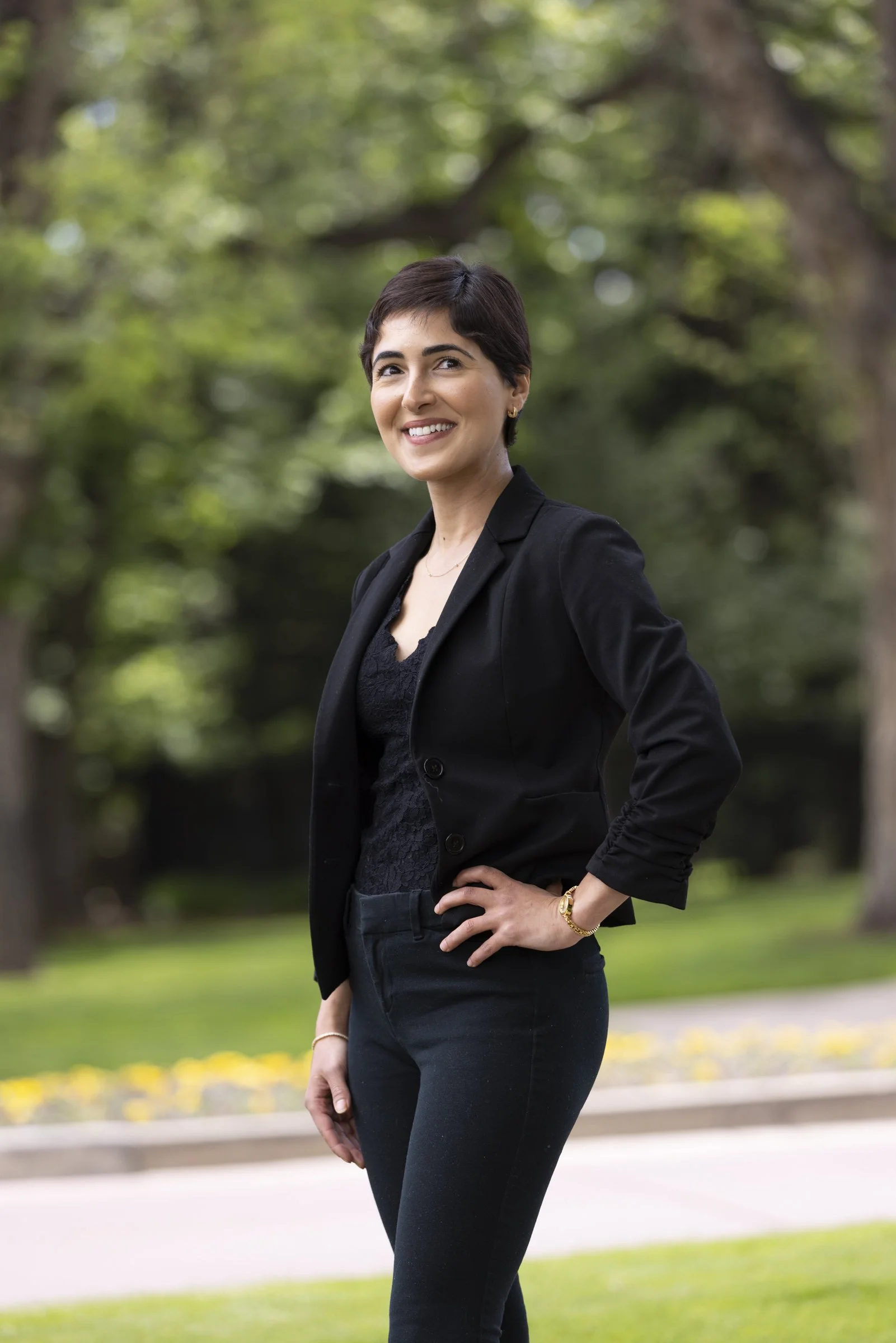 A woman with short dark hair wearing a black blazer and matching black pants stands outdoors in a park with trees and greenery in the background. She is smiling with one hand on her hip.
