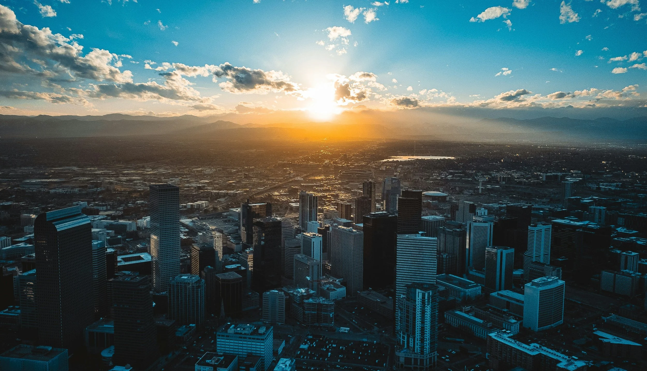City skyline with tall skyscrapers at sunset, mountains in the background, and a partly cloudy sky.