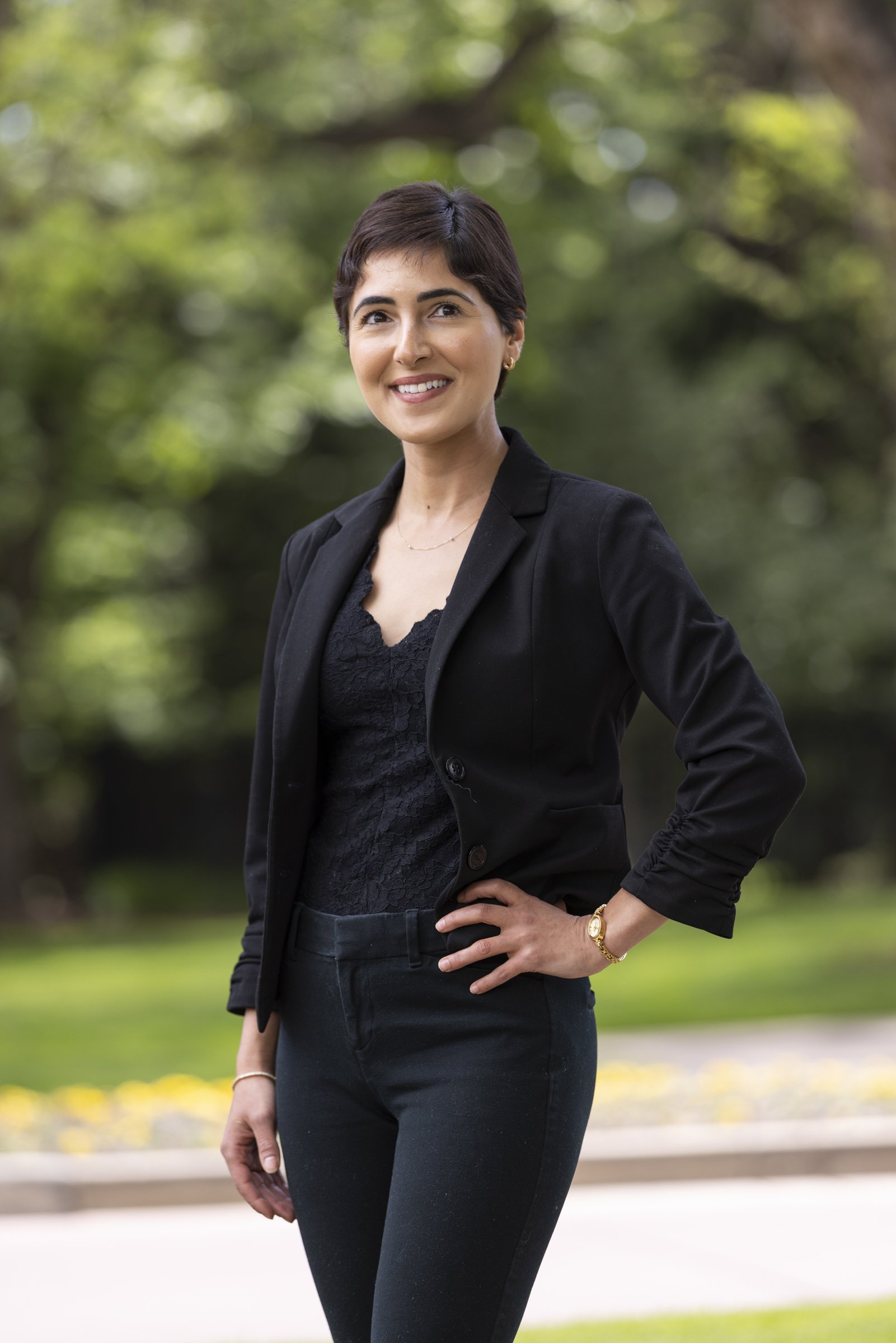 A woman with short dark hair, wearing a black blazer and matching black pants, standing outdoors in a park, smiling, with a blurred green tree background.