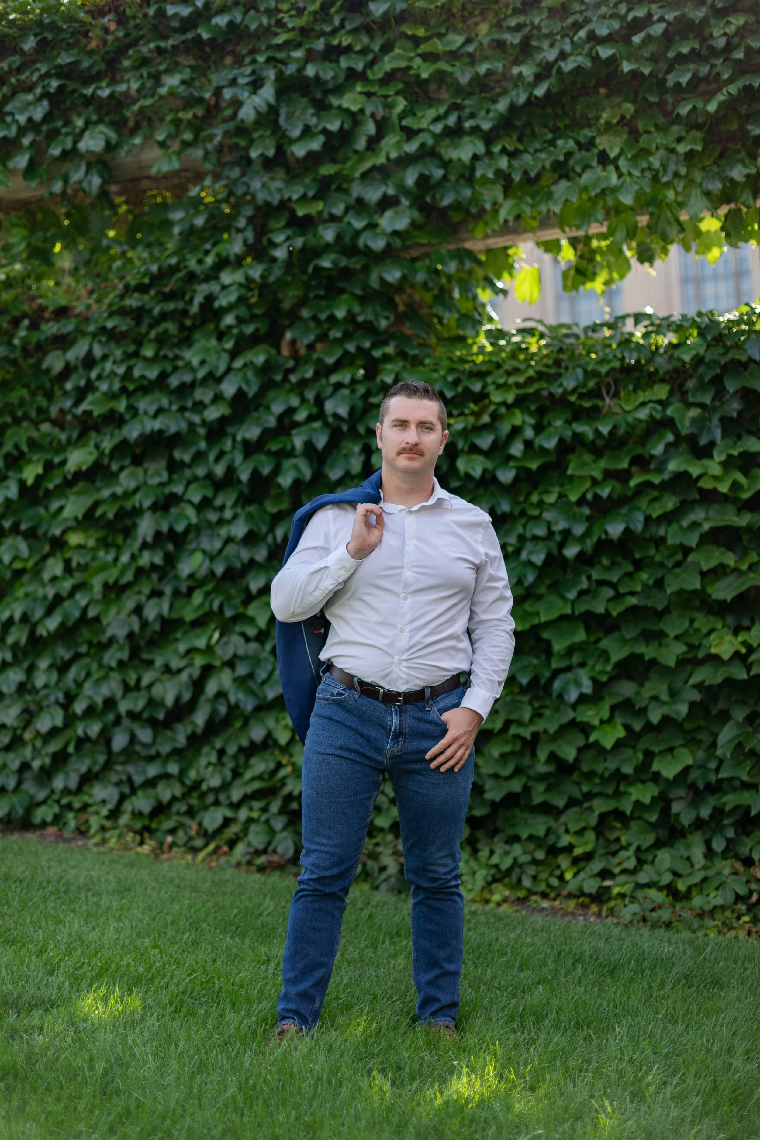 Man standing outdoors on green grass in front of a large wall of ivy, wearing a white shirt and holding a blue jacket over his shoulder.