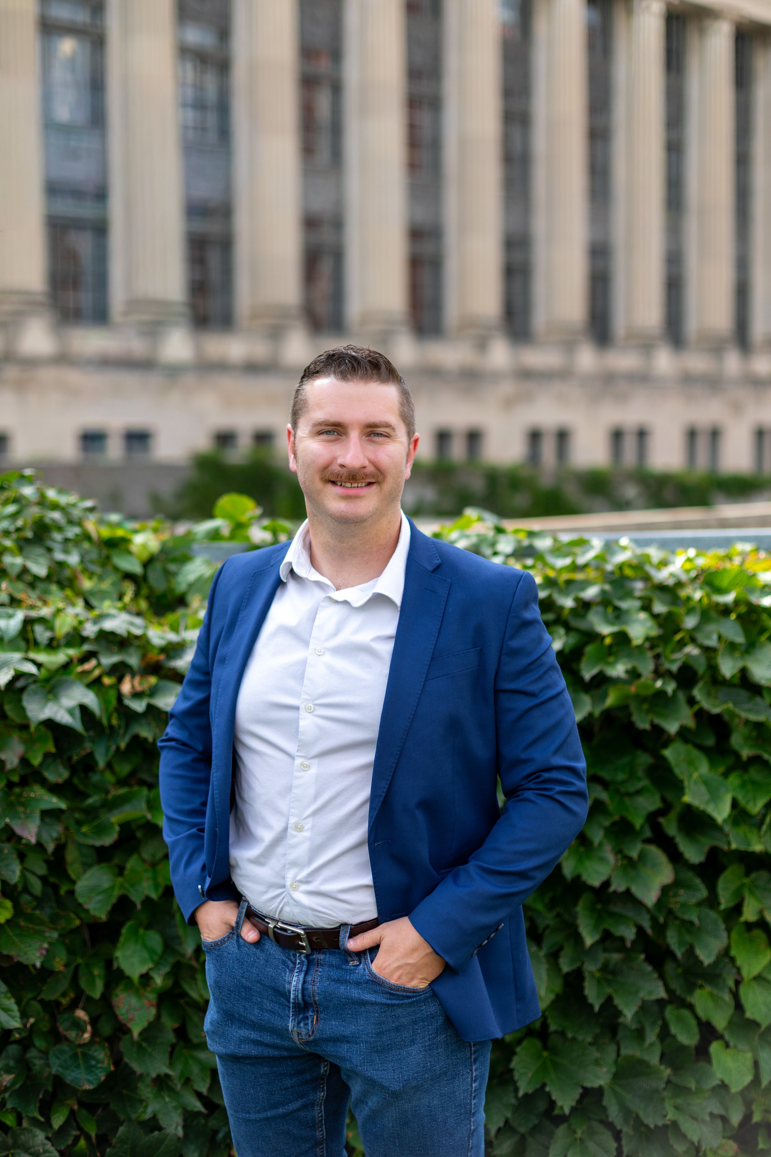 A young man in a blue blazer and jeans smiling outdoors in front of green leafy bushes with a building in the background.