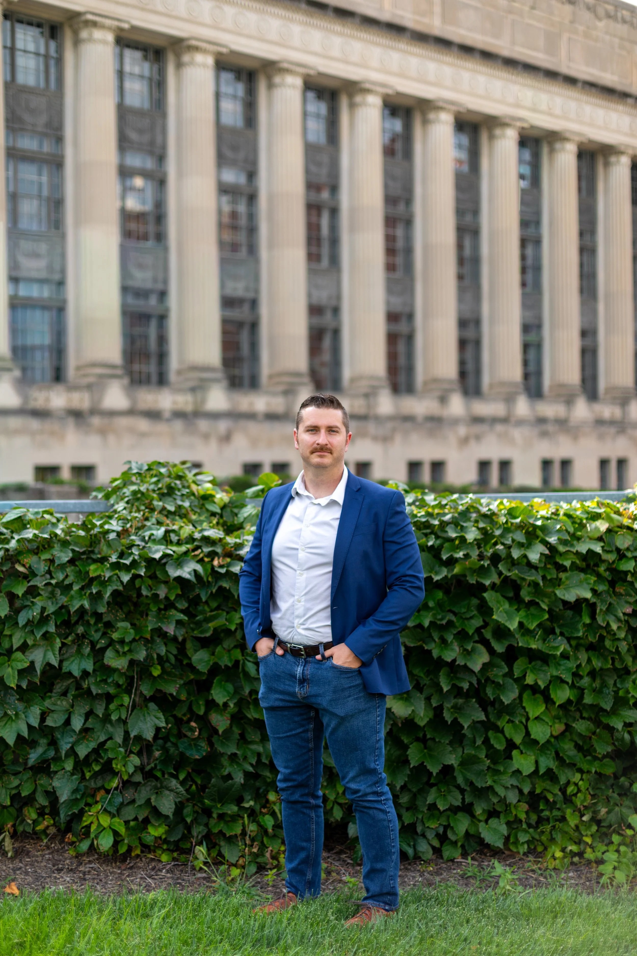 A man in a blue blazer and jeans stands with one hand in his pocket in front of green bushes, with a large, classical-style building with tall columns in the background.