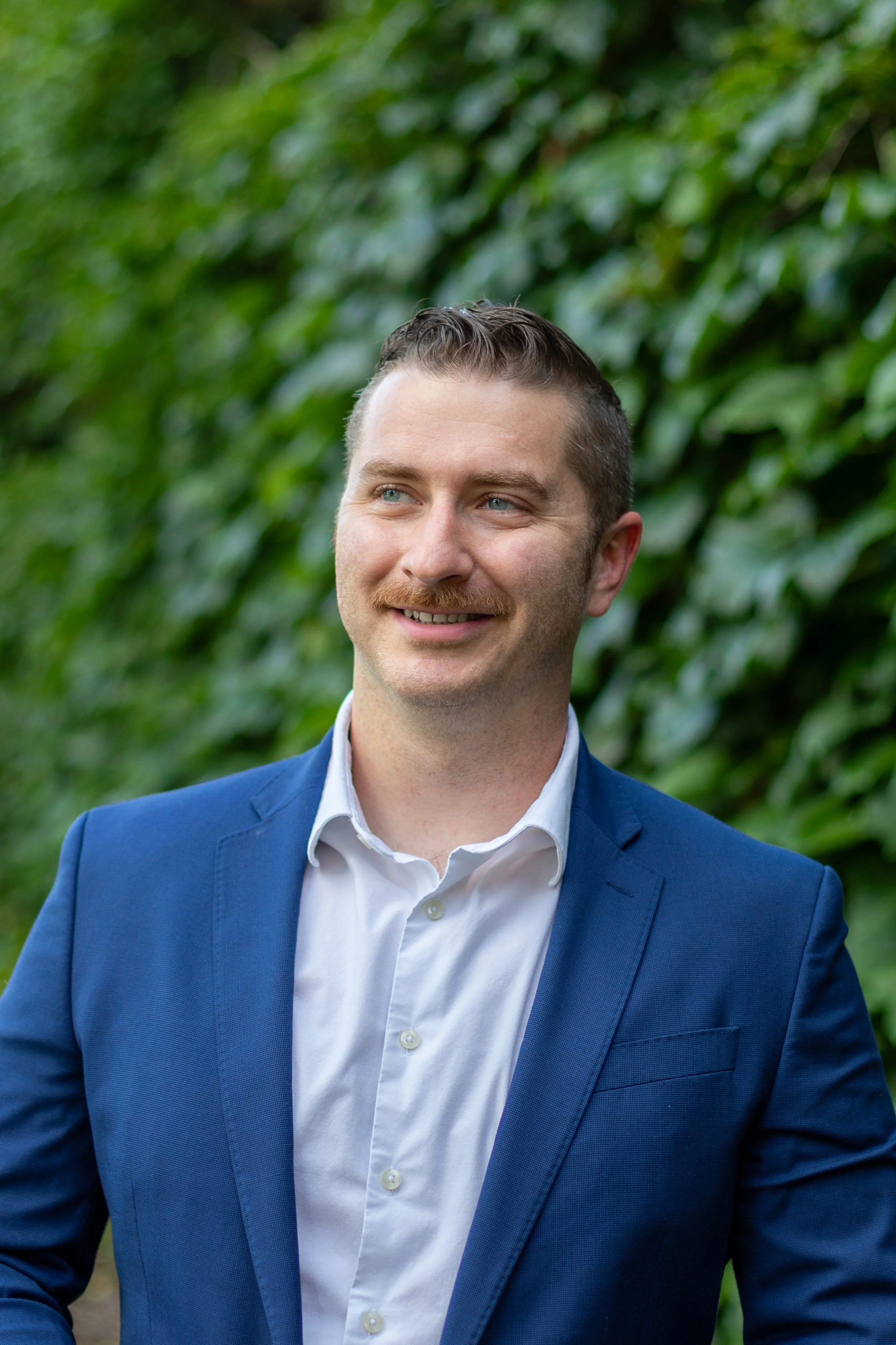A man with light skin, blue eyes, short dark hair, and a mustache, wearing a blue suit jacket and white shirt, smiling outdoors with greenery in the background.
