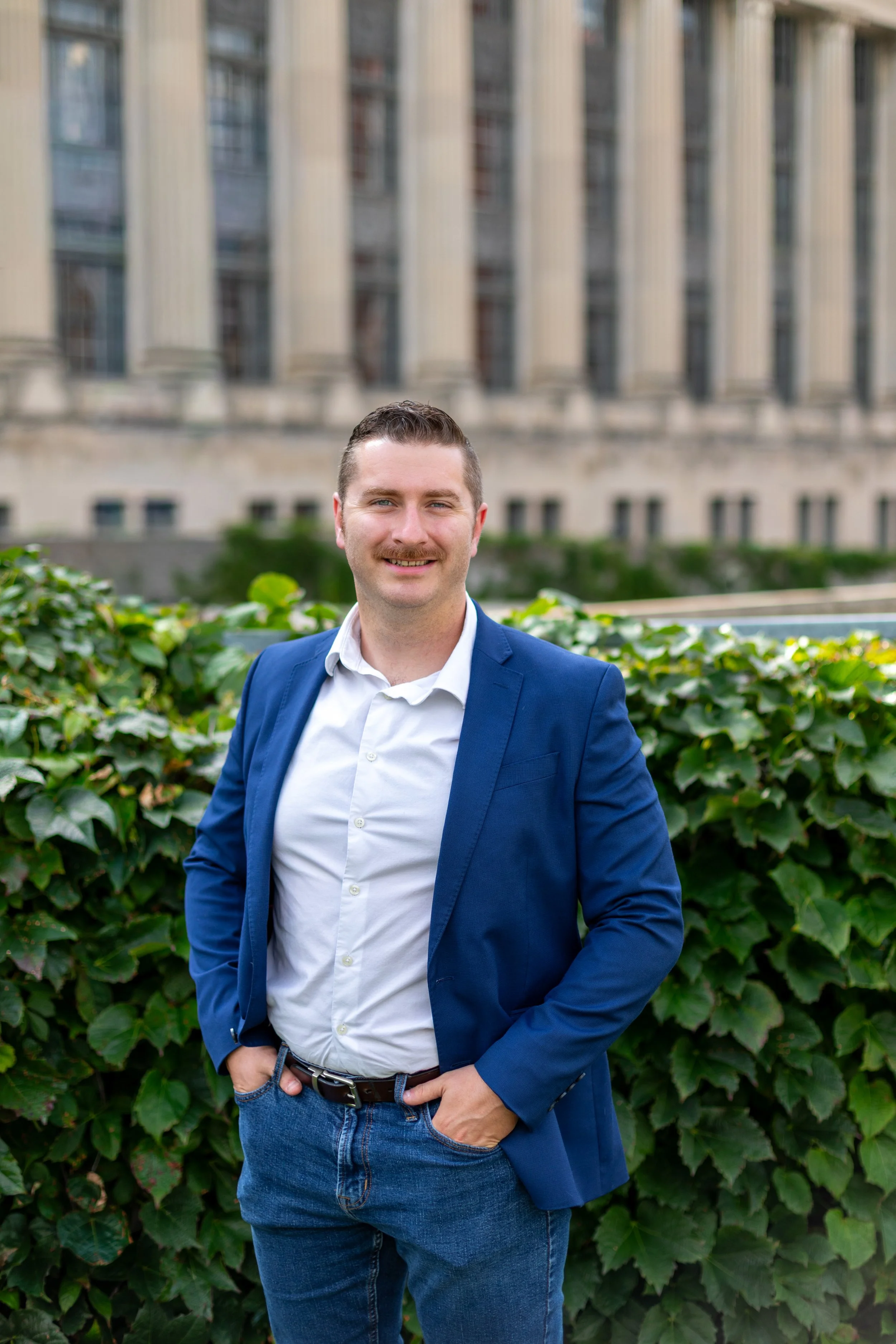 A smiling man in a blue blazer and white shirt standing outdoors in front of a leafy green hedge with a large building in the background.