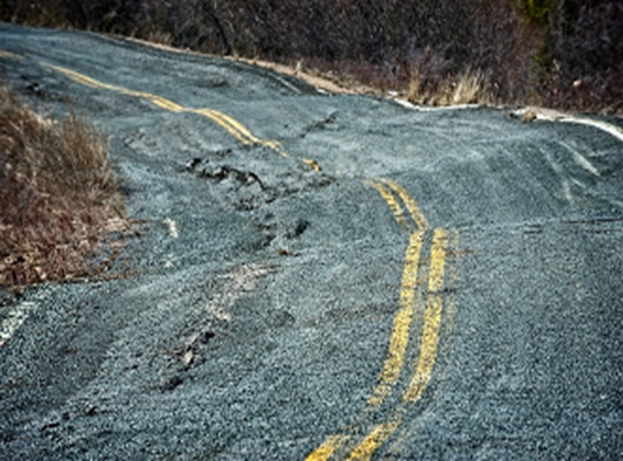 A damaged and cracked road with uneven surface and faded yellow lane markings.