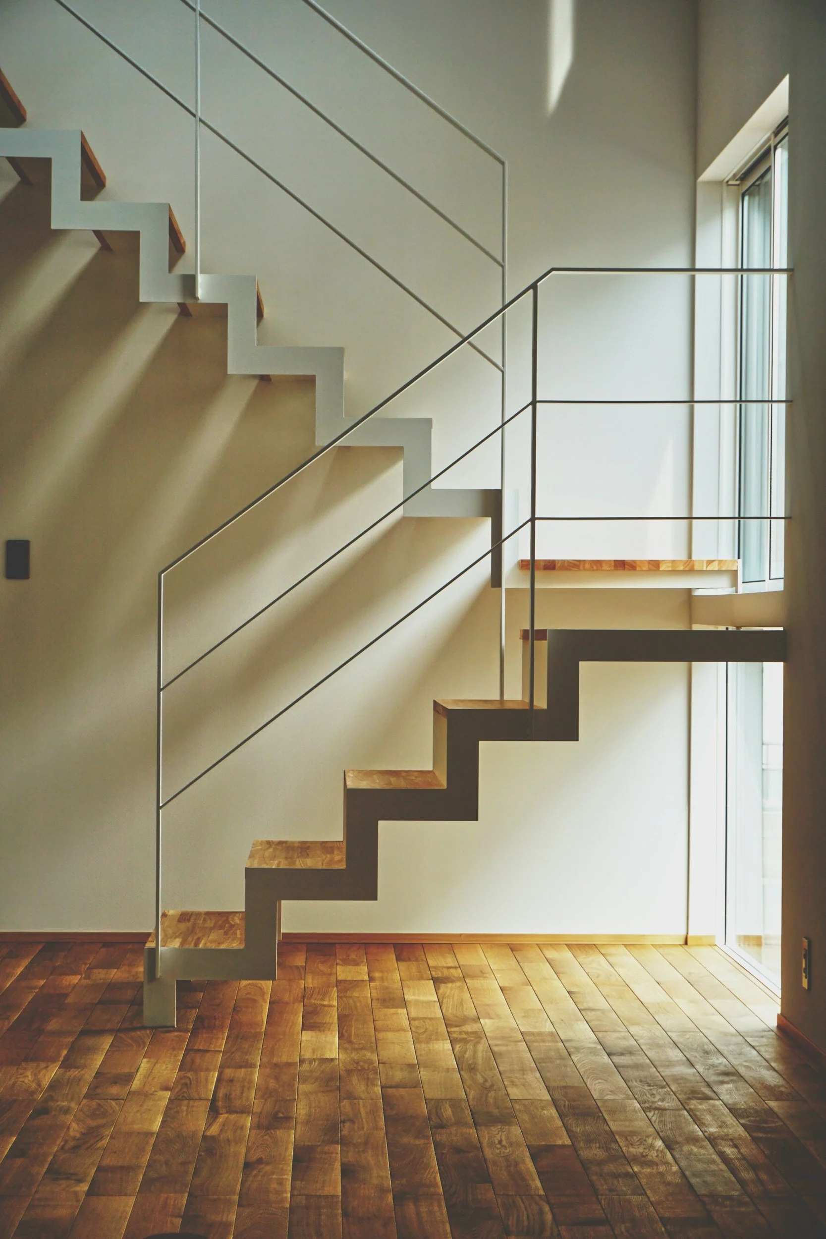 Modern staircase with zigzag pattern, white frame, wooden steps, thin metal rails, next to a large window with sunlight, hardwood floor, beige wall.