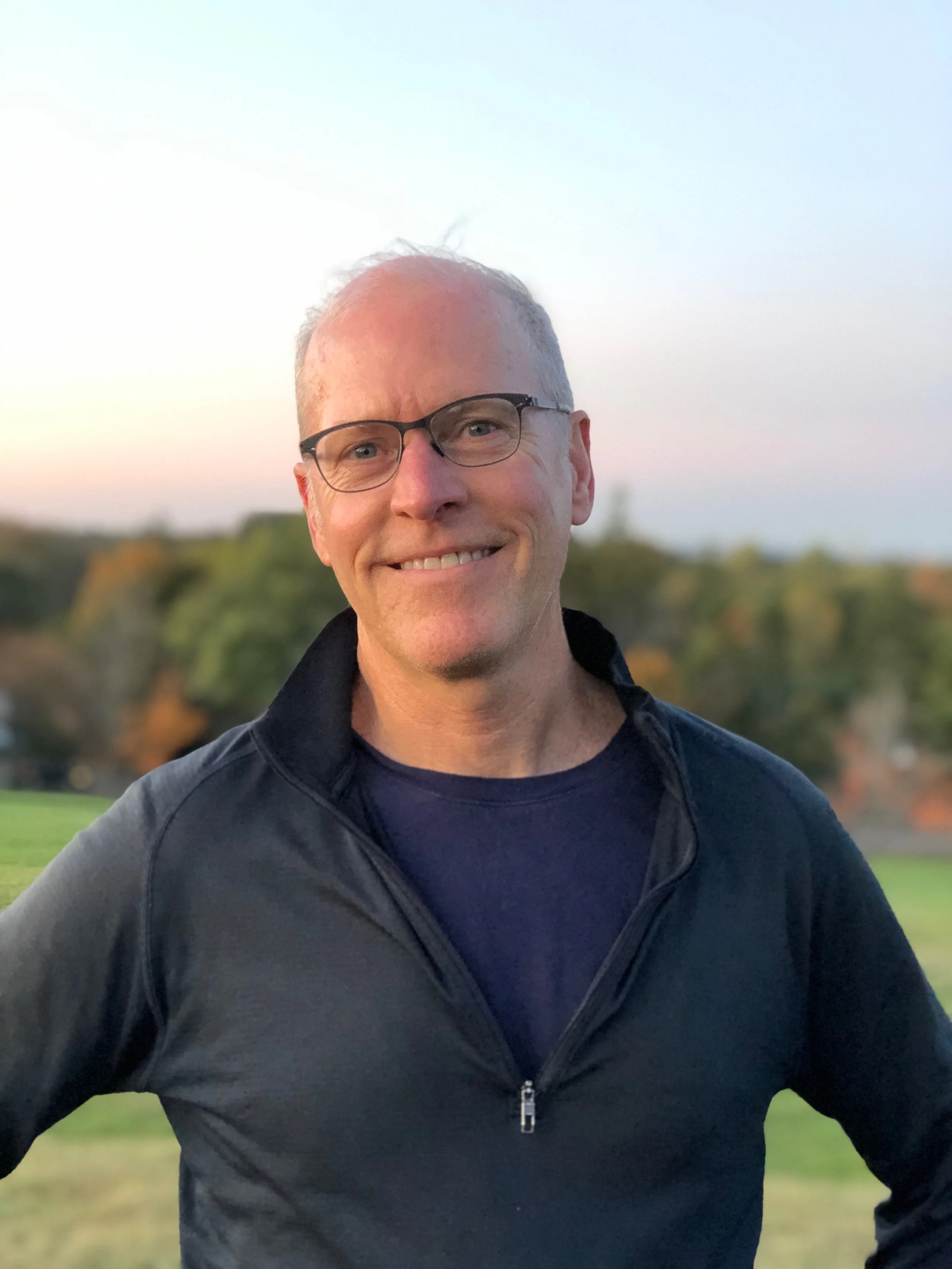 A smiling middle-aged man with glasses wearing a black zippered jacket, standing outdoors with a blurred background of green trees and a pastel sky during sunset.