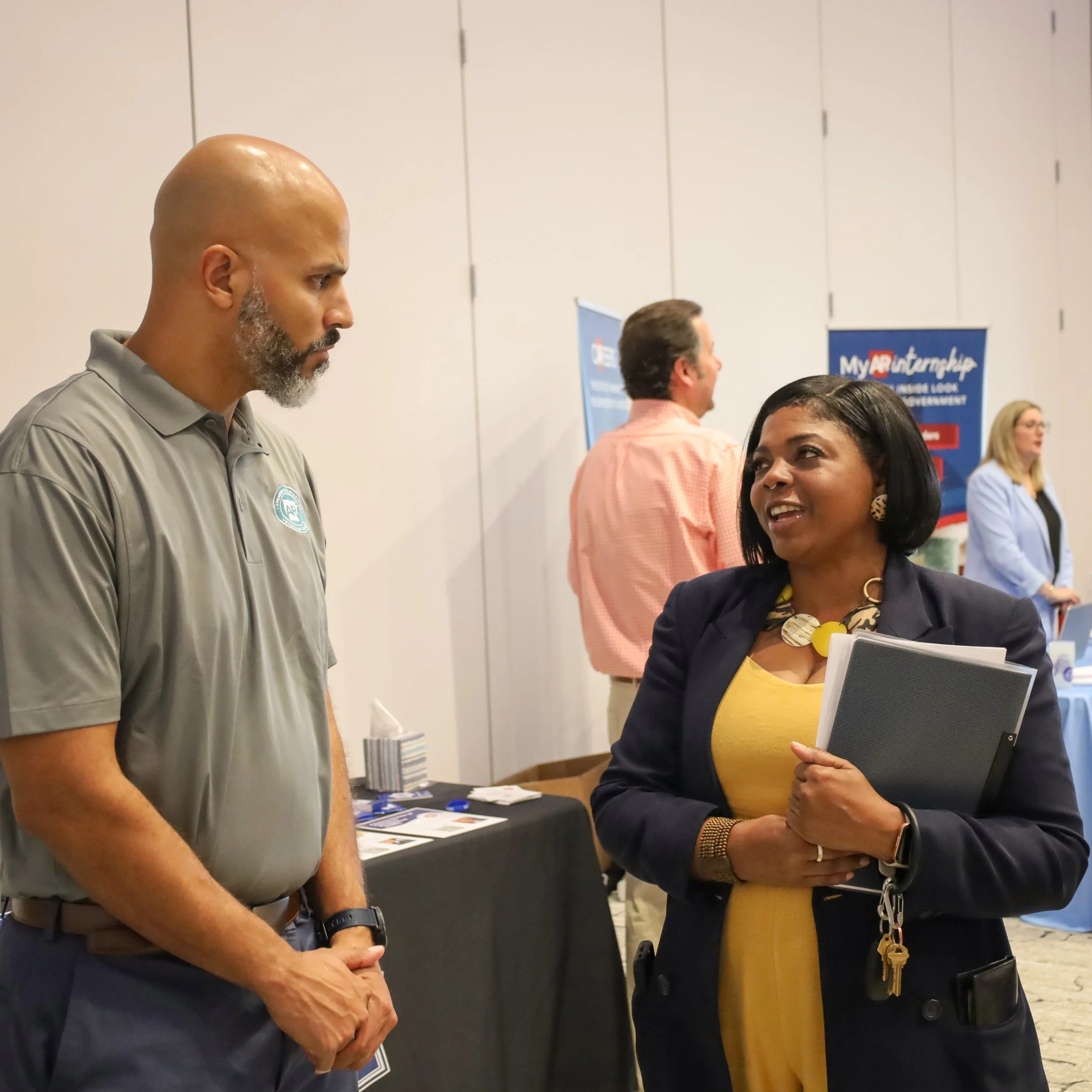 Two professionals converse at a networking event, with promotional booths and attendees in the background.