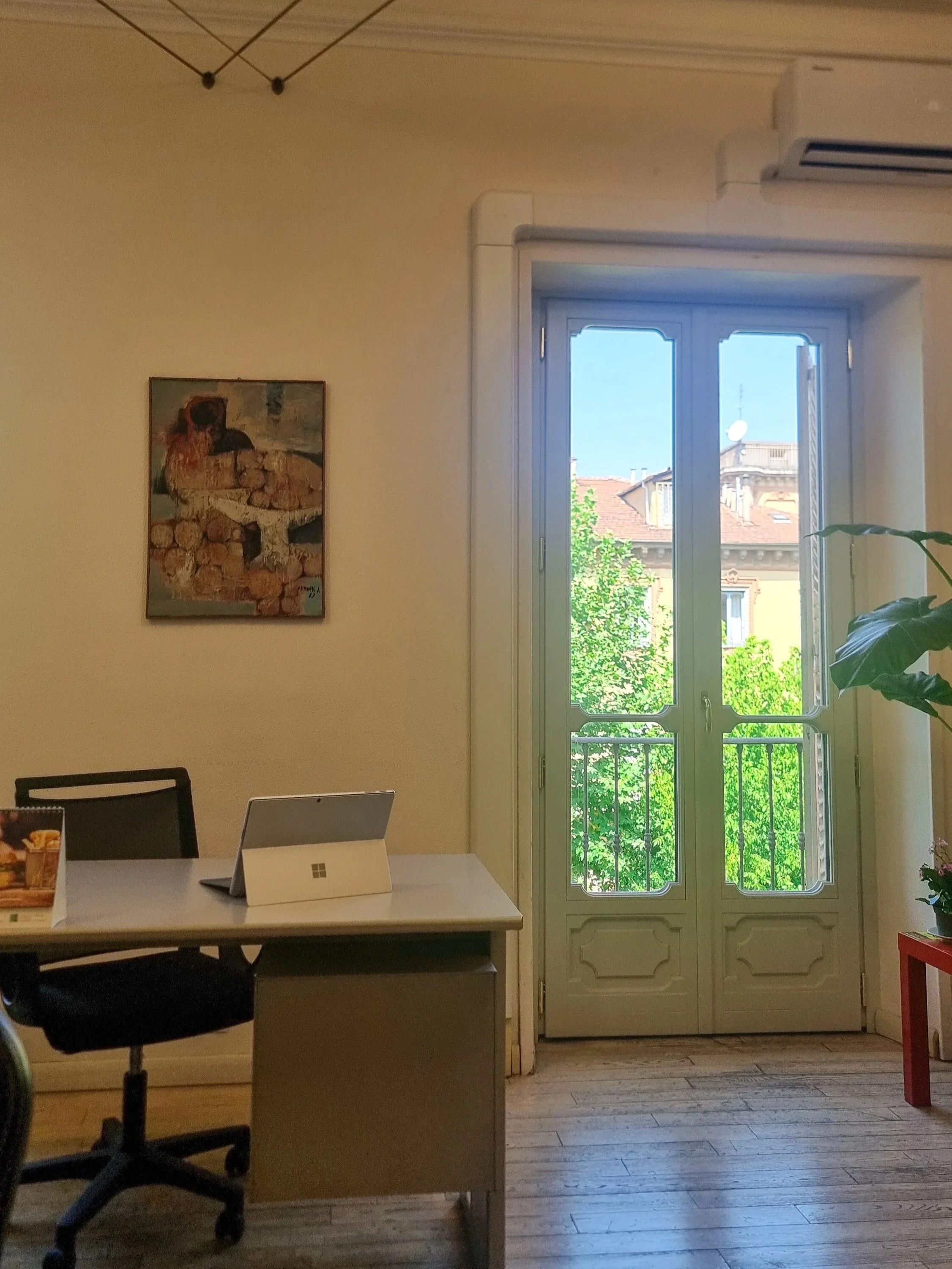 Ufficio con una scrivania, una sedia con rotelle, una finestra grande con balcone e vista sulla città, un quadro appeso alla parete e una pianta