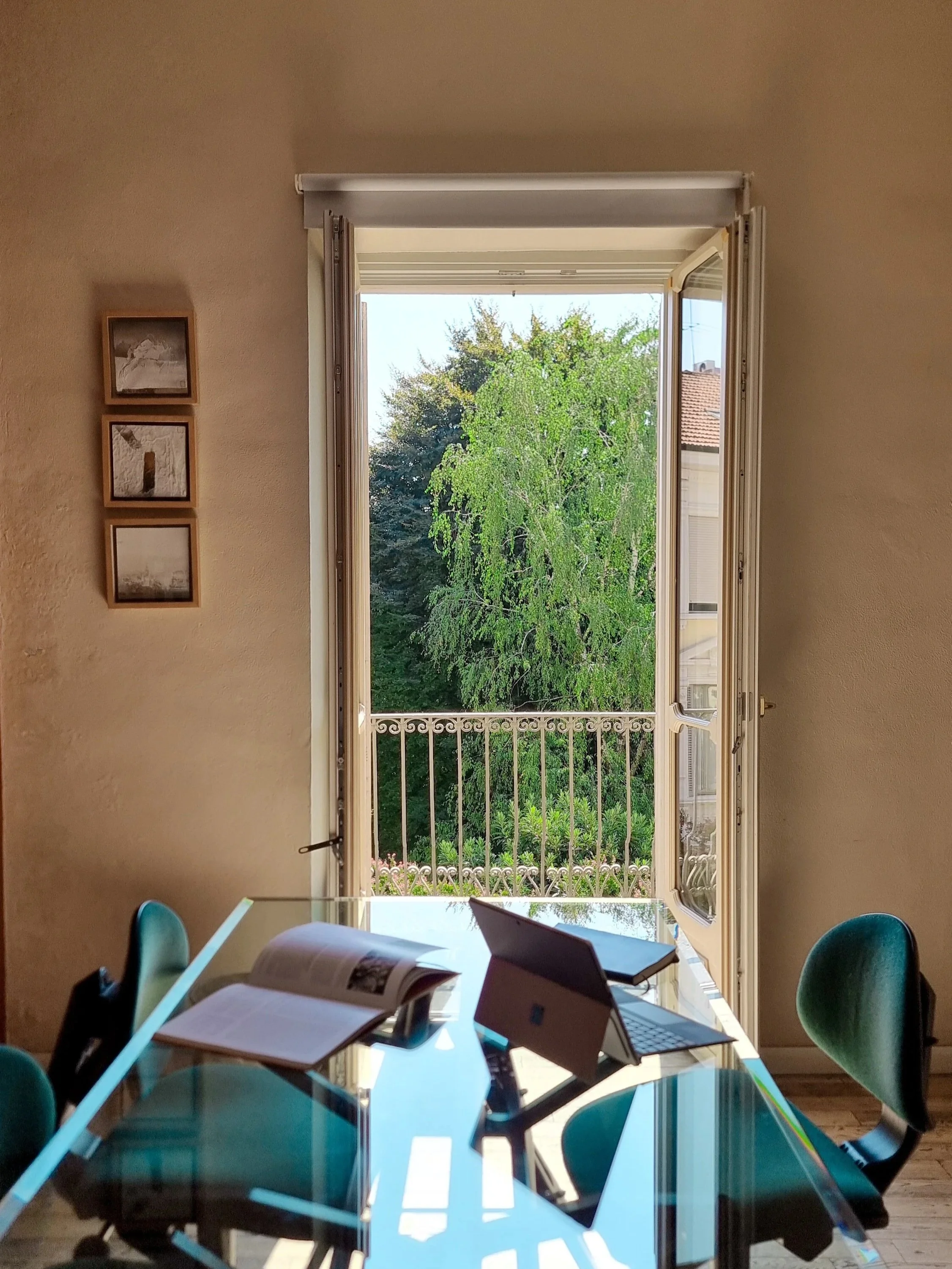 Interno di una stanza con una grande finestra aperta, che mostra alberi verdi e un tetto di una casa. Sul tavolo ci sono libri, un laptop, un tablet, una tazza e un computer portatile. Sui sedie ci sono due persone sedute, una di spalle e l'altra di fronte con il viso non visibile.