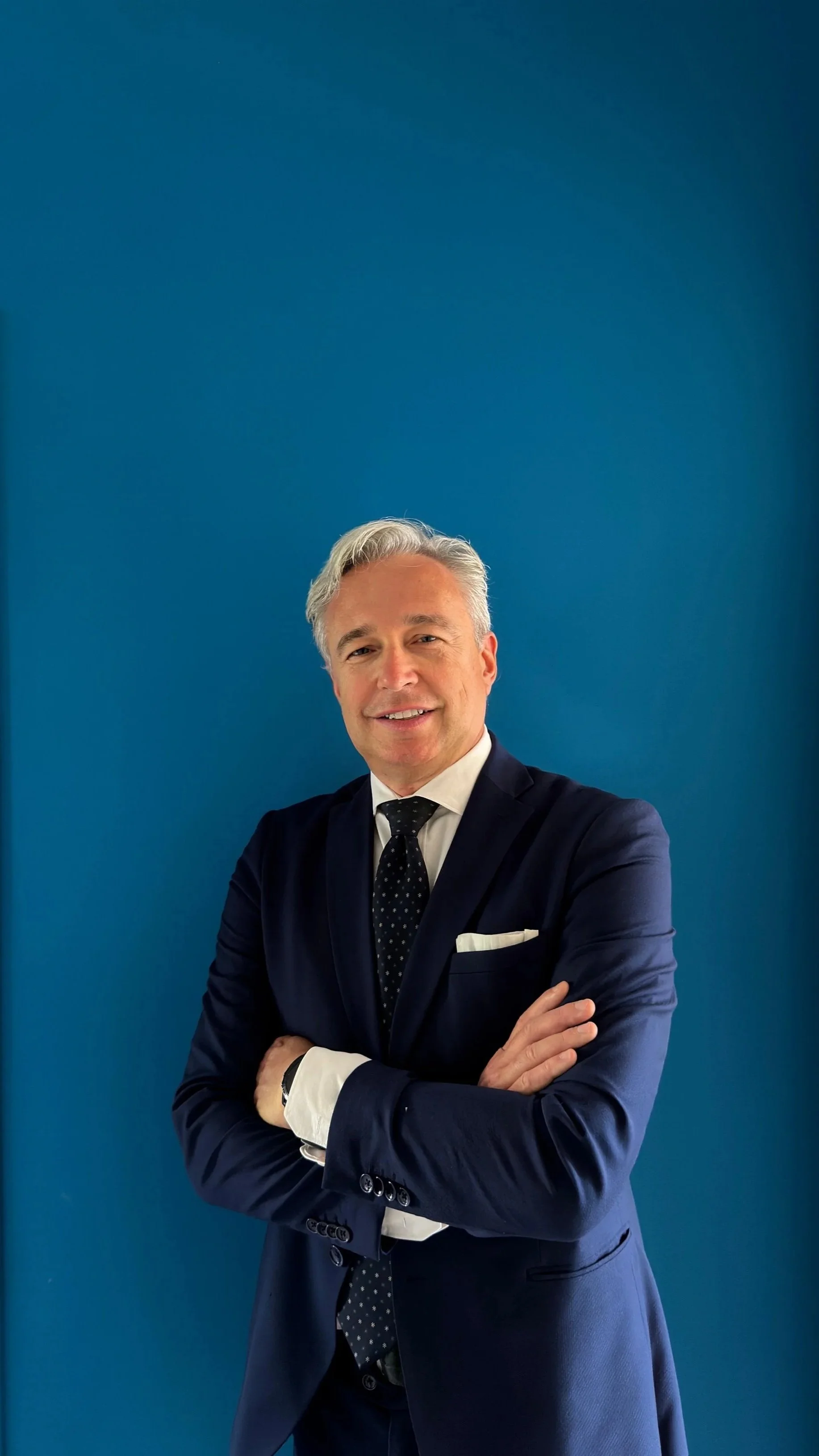 Uomo d'affari in abito blu scuro con cravatta a pois, con le braccia incrociate, sorridente, davanti a uno sfondo blu
