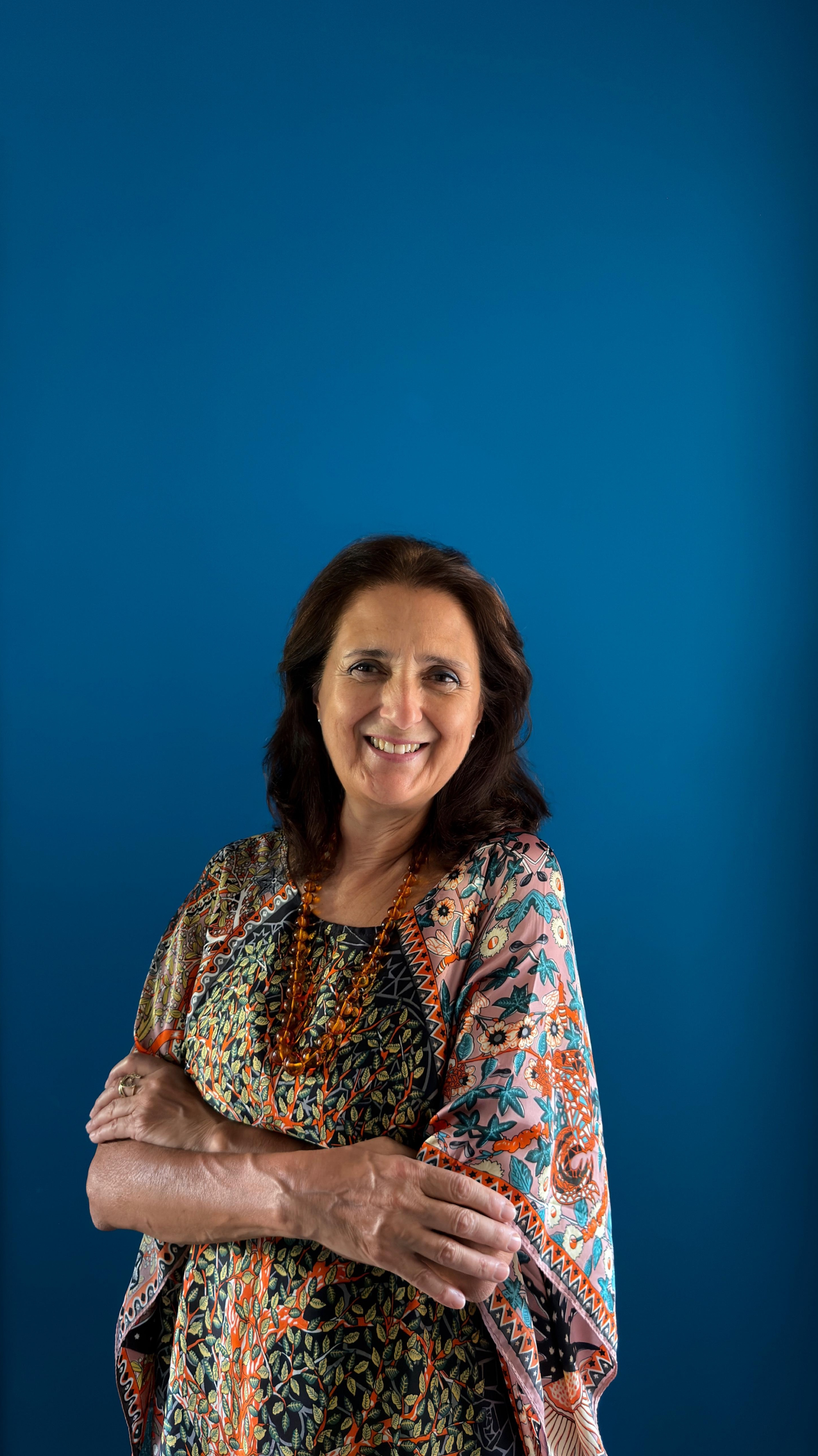 Una donna sorridente con capelli castani e camicia con motivi floreali e geometrici, indossa collana di bigiotteria, con braccia incrociate, sfondo blu.