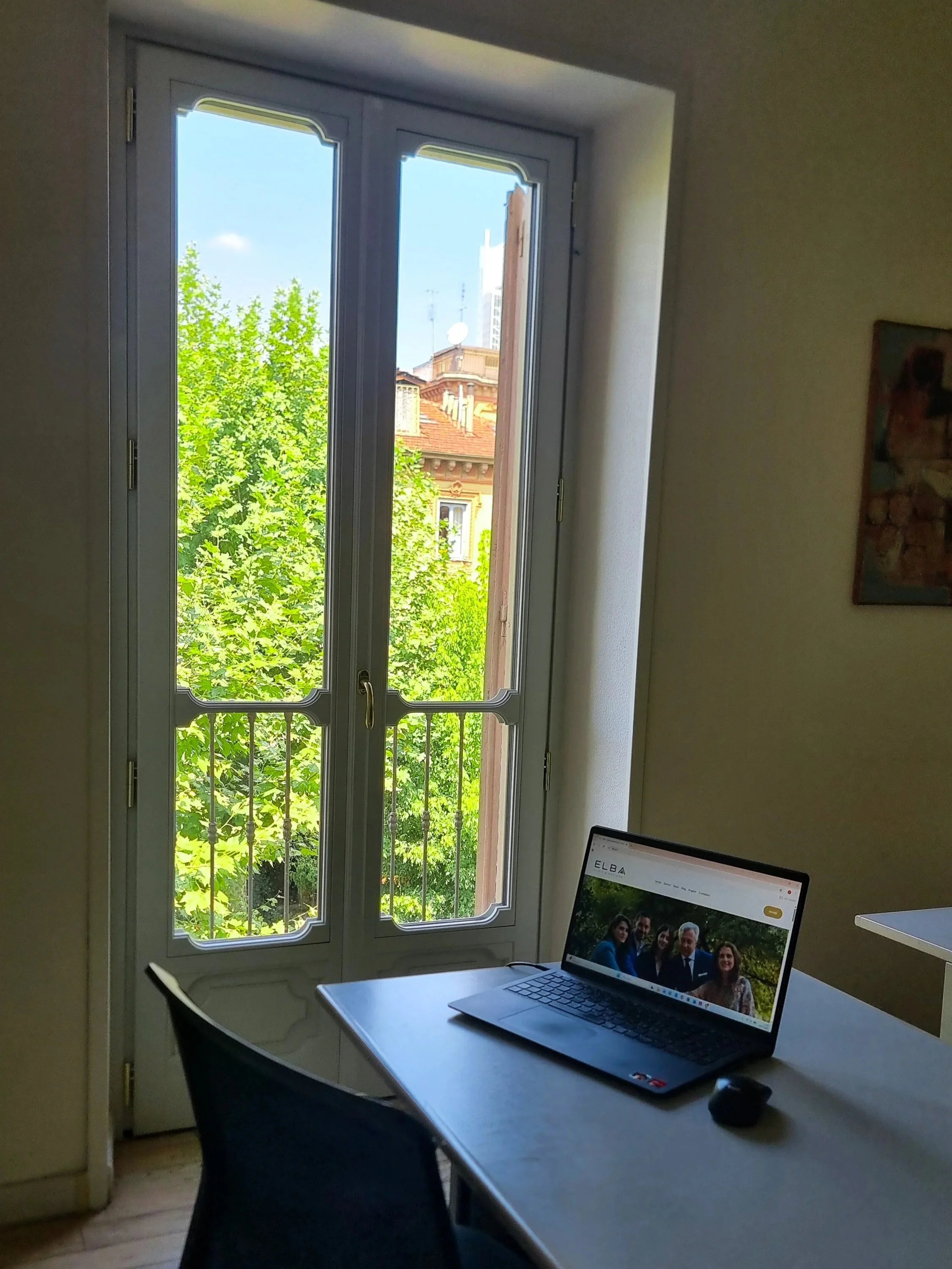 Una scrivania con un computer portatile e una sedia, vicino a una grande finestra che dà su un albero verde e un edificio con tetto rosso e comignoli.