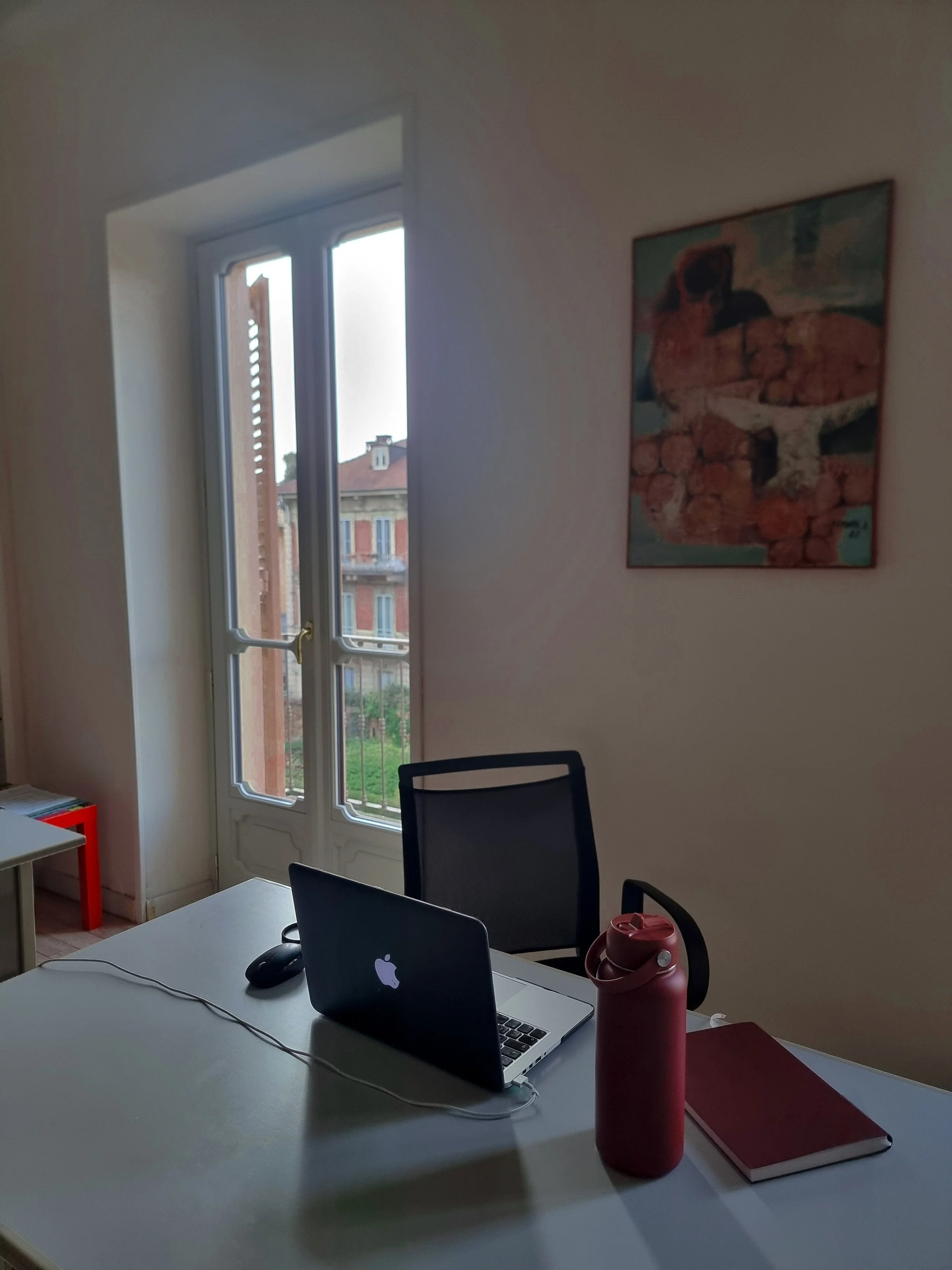 Ufficio con finestra che dà su edifici. Tavolo con un laptop, un mouse e una bottiglia di tè. Sedia, libro e poster alla parete.
