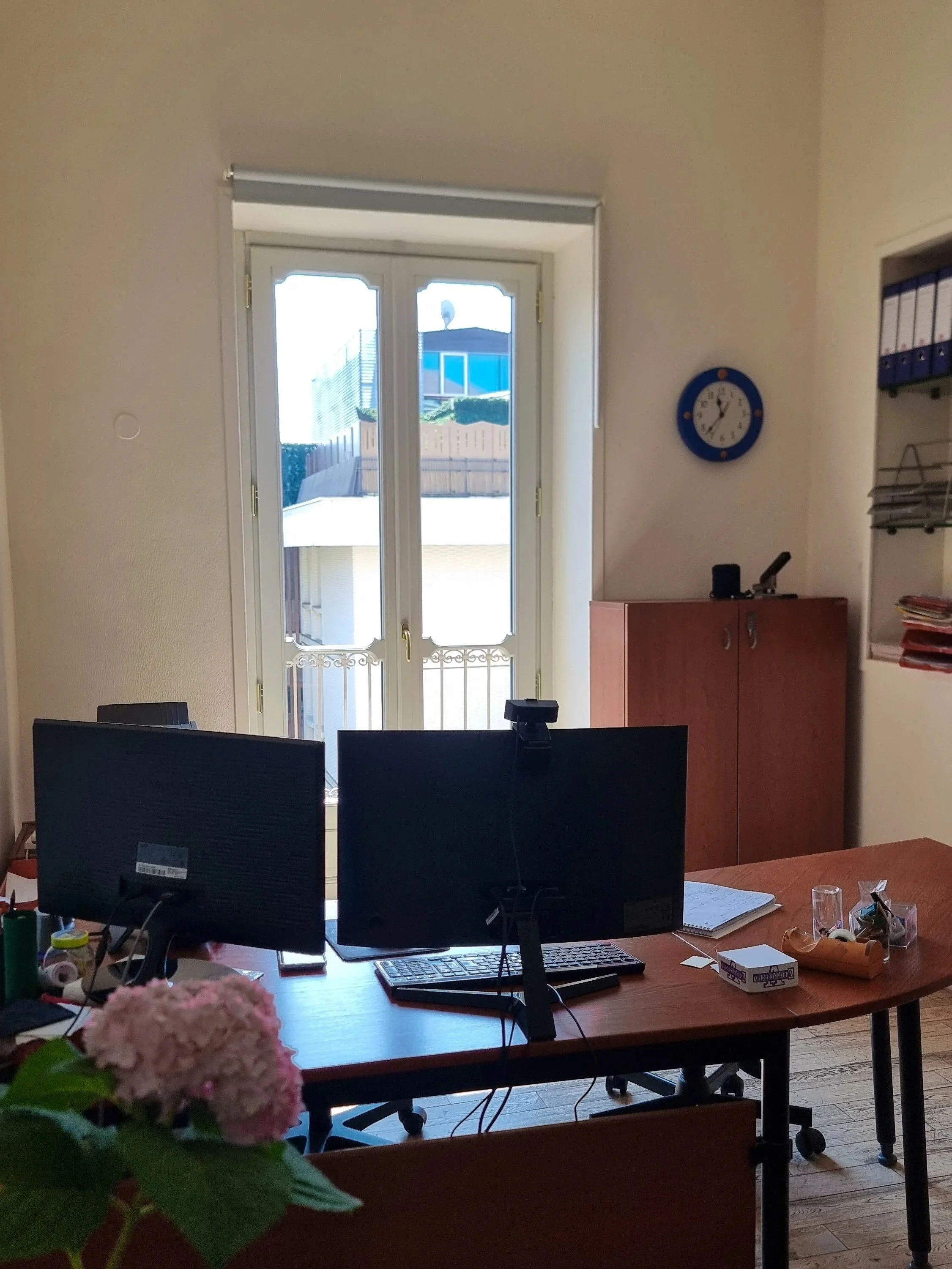 Interno di un ufficio con due schermi di computer, una finestra grande che dà sulla strada con un giardino e un edificio, orologio sulla parete, lasciando intravedere vari oggetti da scrivania e alcuni file.