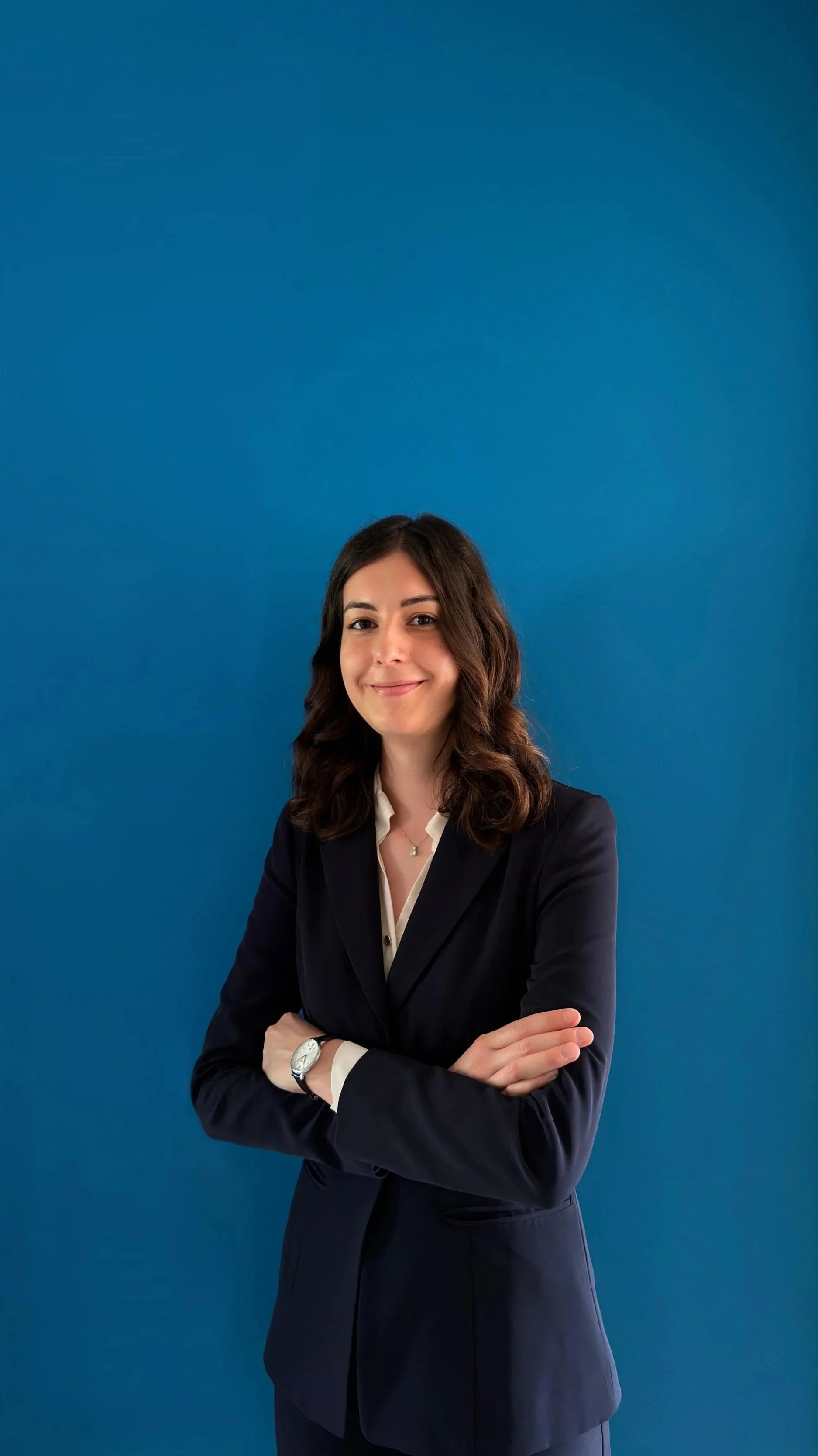 Una donna sorridente con capelli castani ondulati, vestita con abito nero e camicia bianca, con gli incroci delle braccia davanti a uno sfondo blu.