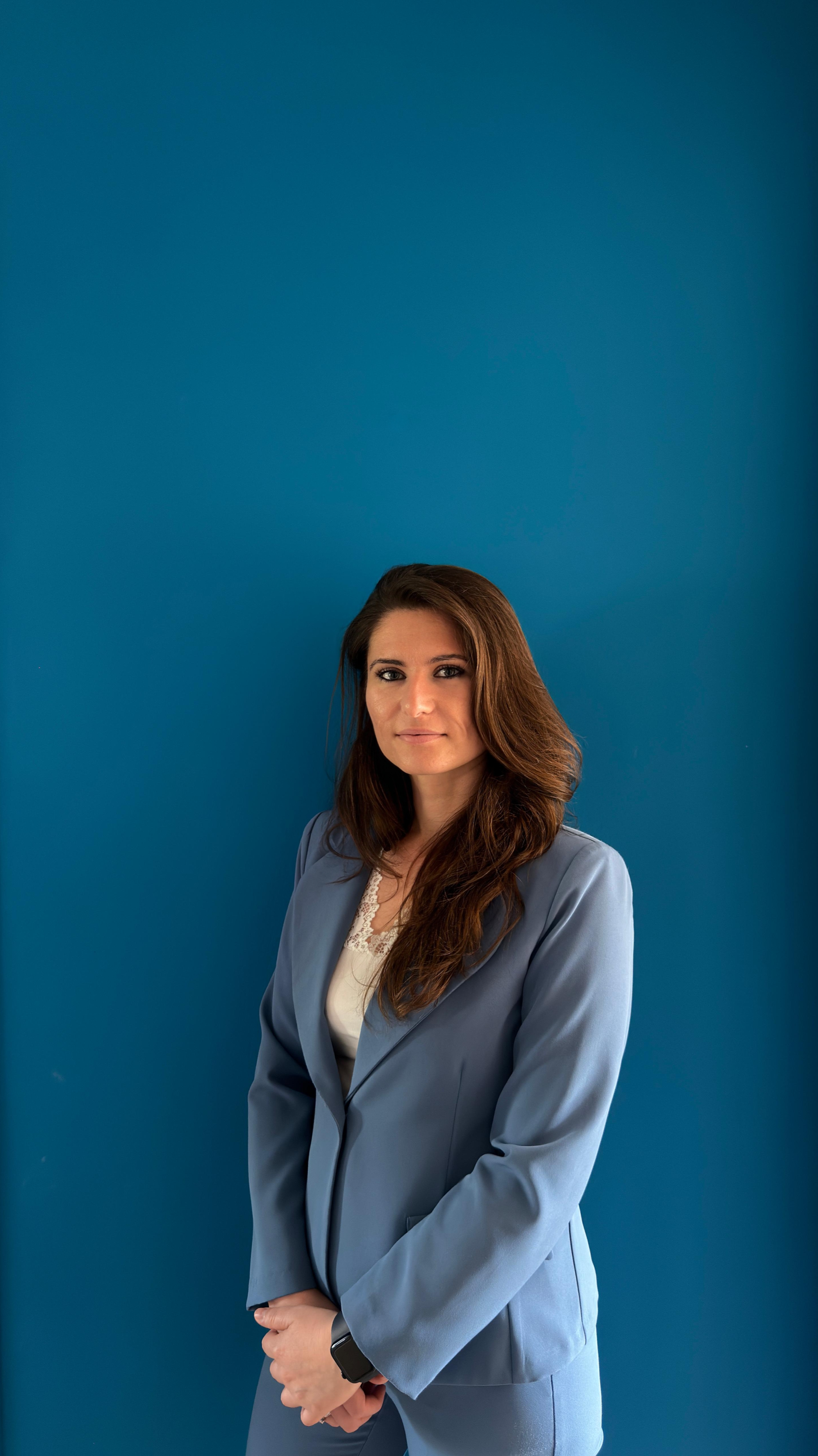 Una donna con capelli castani ricci, vestita con un completo blu, si trova davanti a una parete blu, con uno sguardo serio rivolto verso la telecamera.
