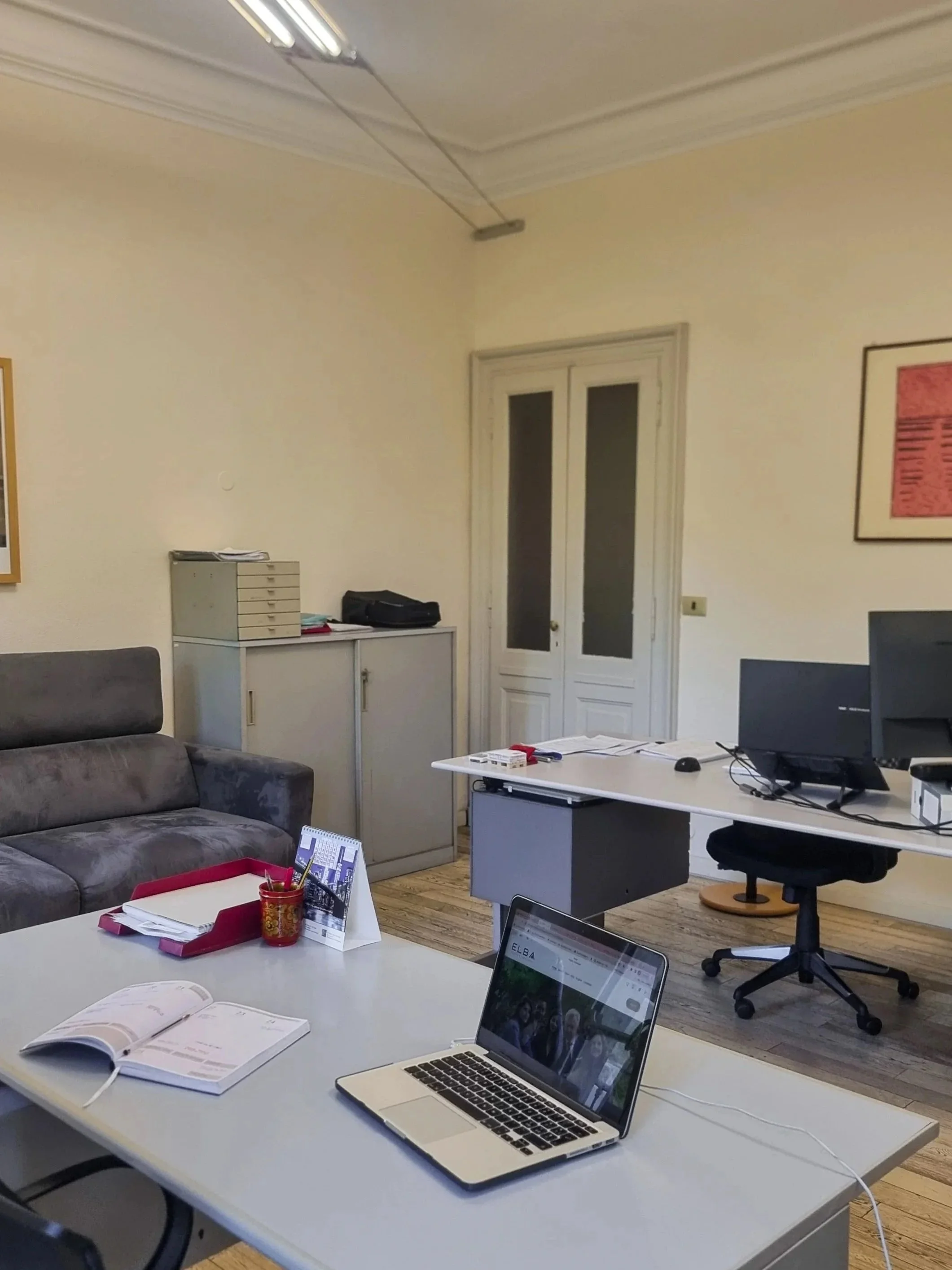 Interno di un ufficio con tavoli, sedie, computer portatile, libri, documenti, e arredamento semplice. Pareti chiare e una porta finestra.