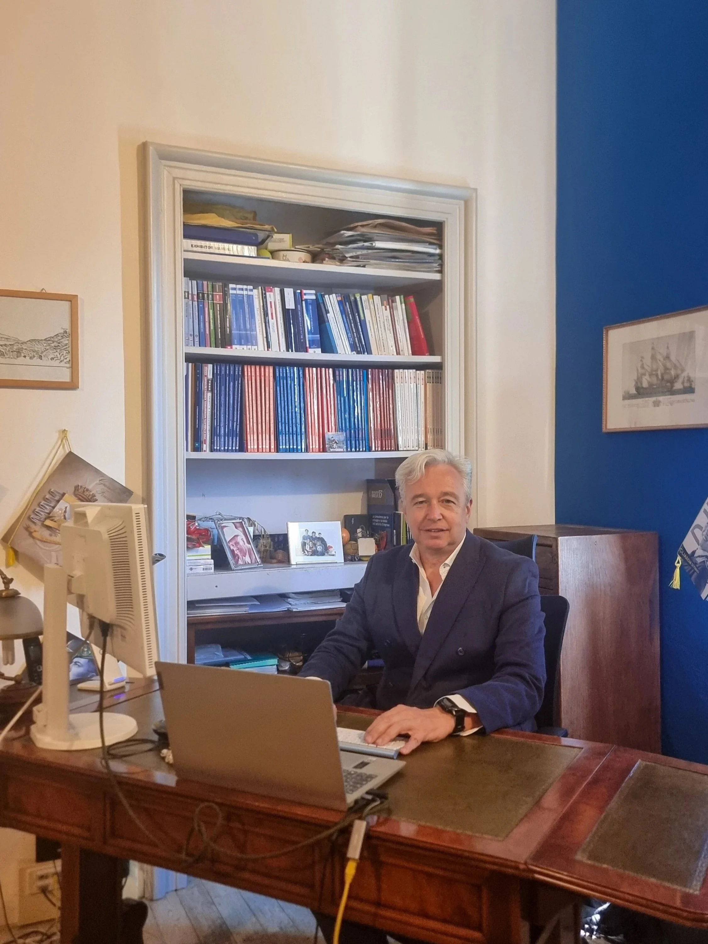 Uomo seduto dietro una scrivania con un laptop, un computer a monitor e una pila di documenti, in un ufficio con scaffali pieni di libri e quadri alle pareti.