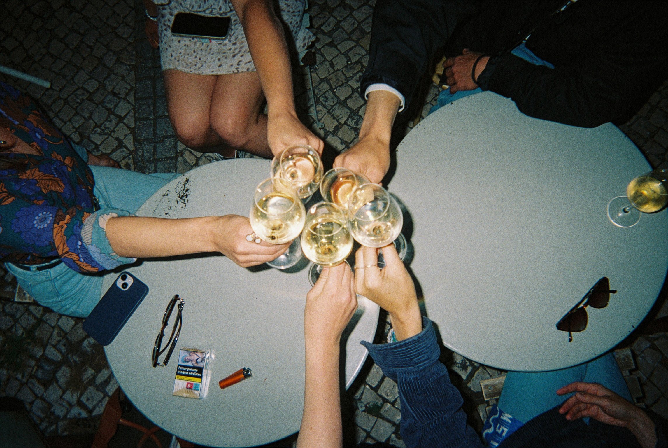 People creating a toast with champagne glasses on a white round table outdoors at night, with sunglasses, a pack of cigarettes, and a phone on the table.