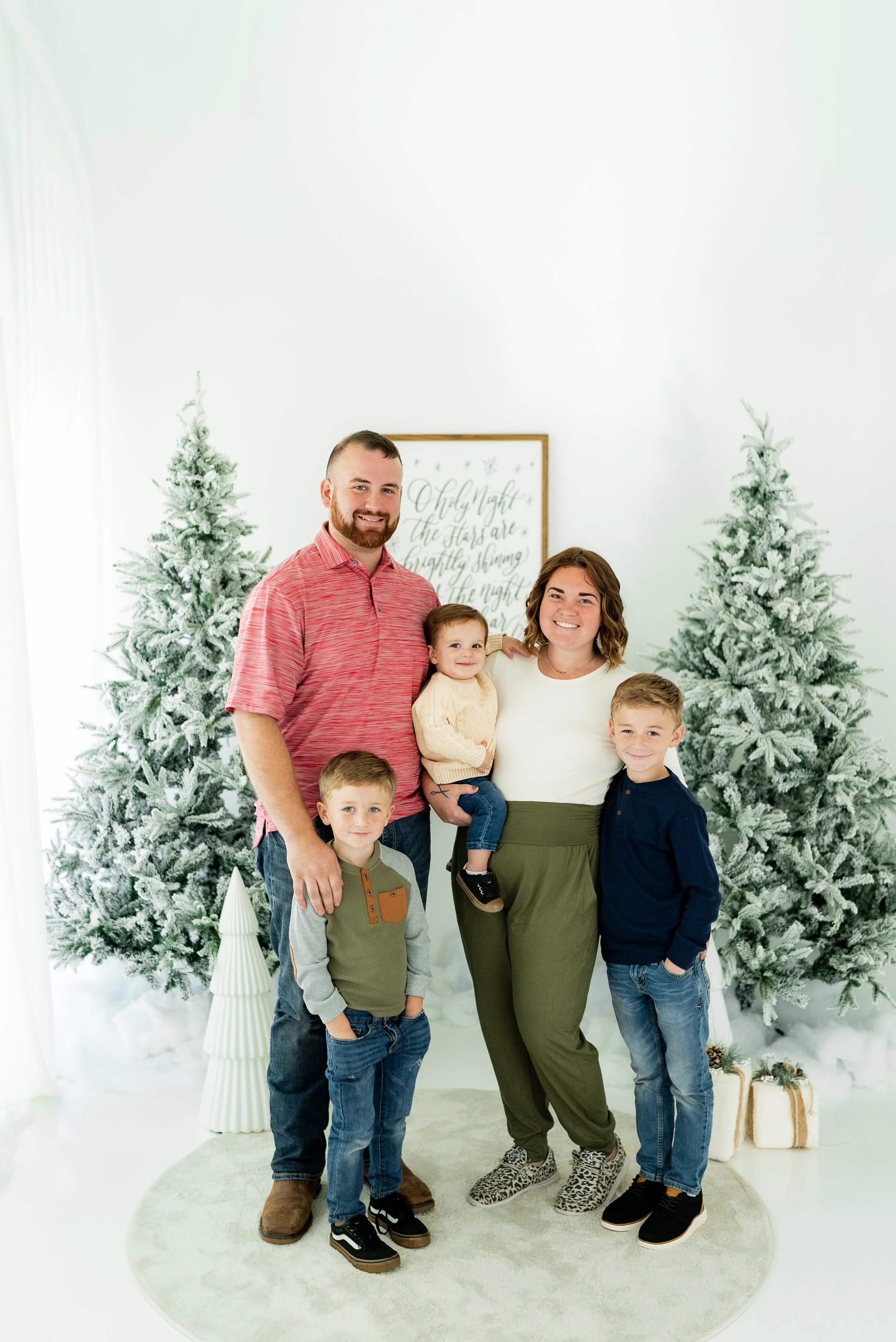 A family of six people smiling and posing in front of decorated snowy Christmas trees and festive decor indoors. A man with a beard in a red shirt, a woman with curly hair and a white shirt, and four children, one in a mustard sweater, one in a blue jacket, and two younger kids, one being held by the woman. Christmas presents are visible on the floor.