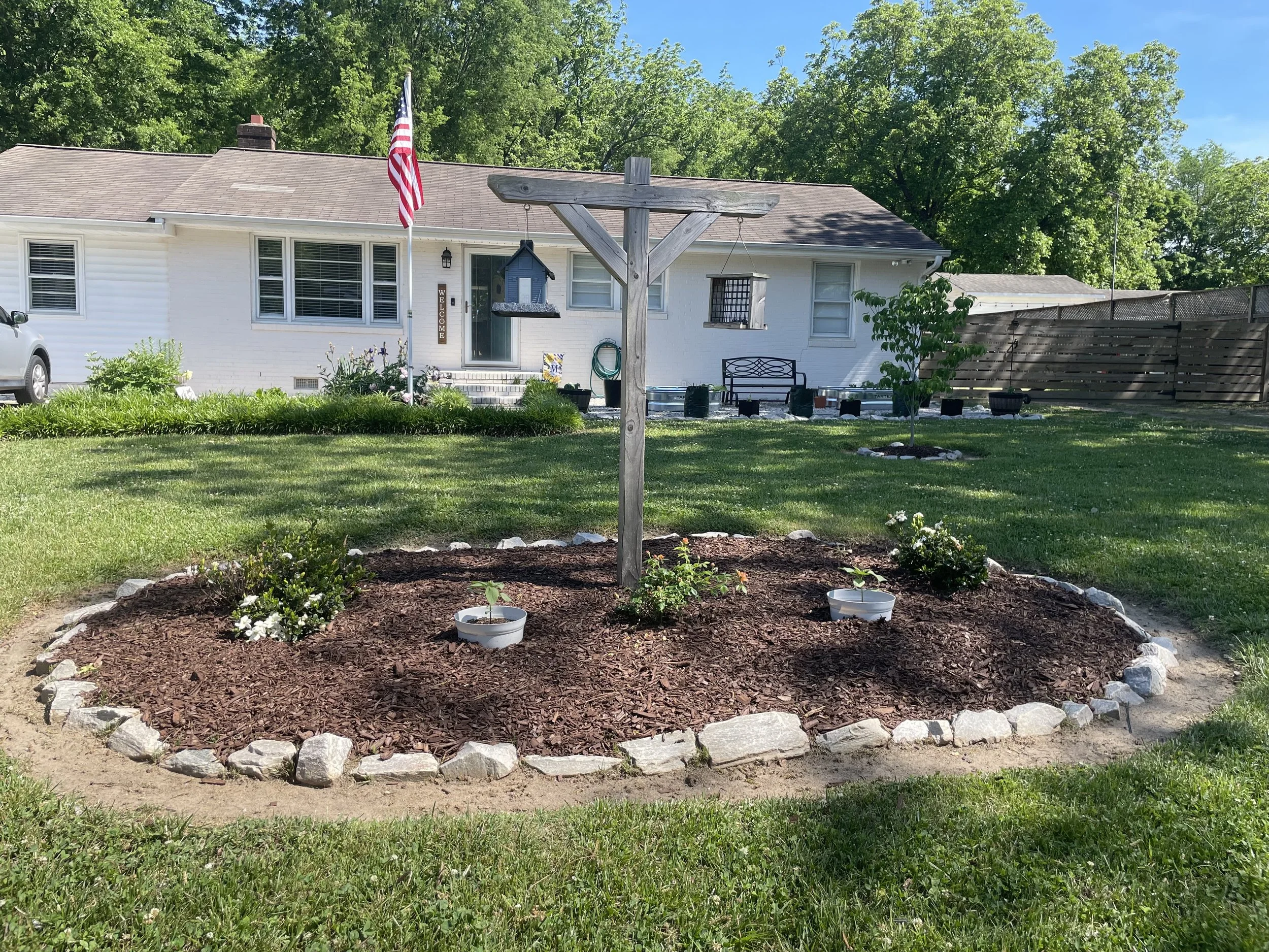 A backyard garden with a flower bed bordered by rocks, plants, and flowers, and a wooden birdhouse on a post. In the background, a white house with a flag, window blinds, and a fence is visible.