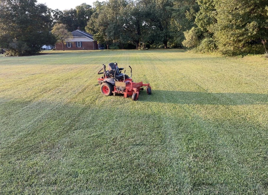 A orange lawn mower on a well-kept grassy field, with trees and a house in the background, during late afternoon or early evening sunlight.