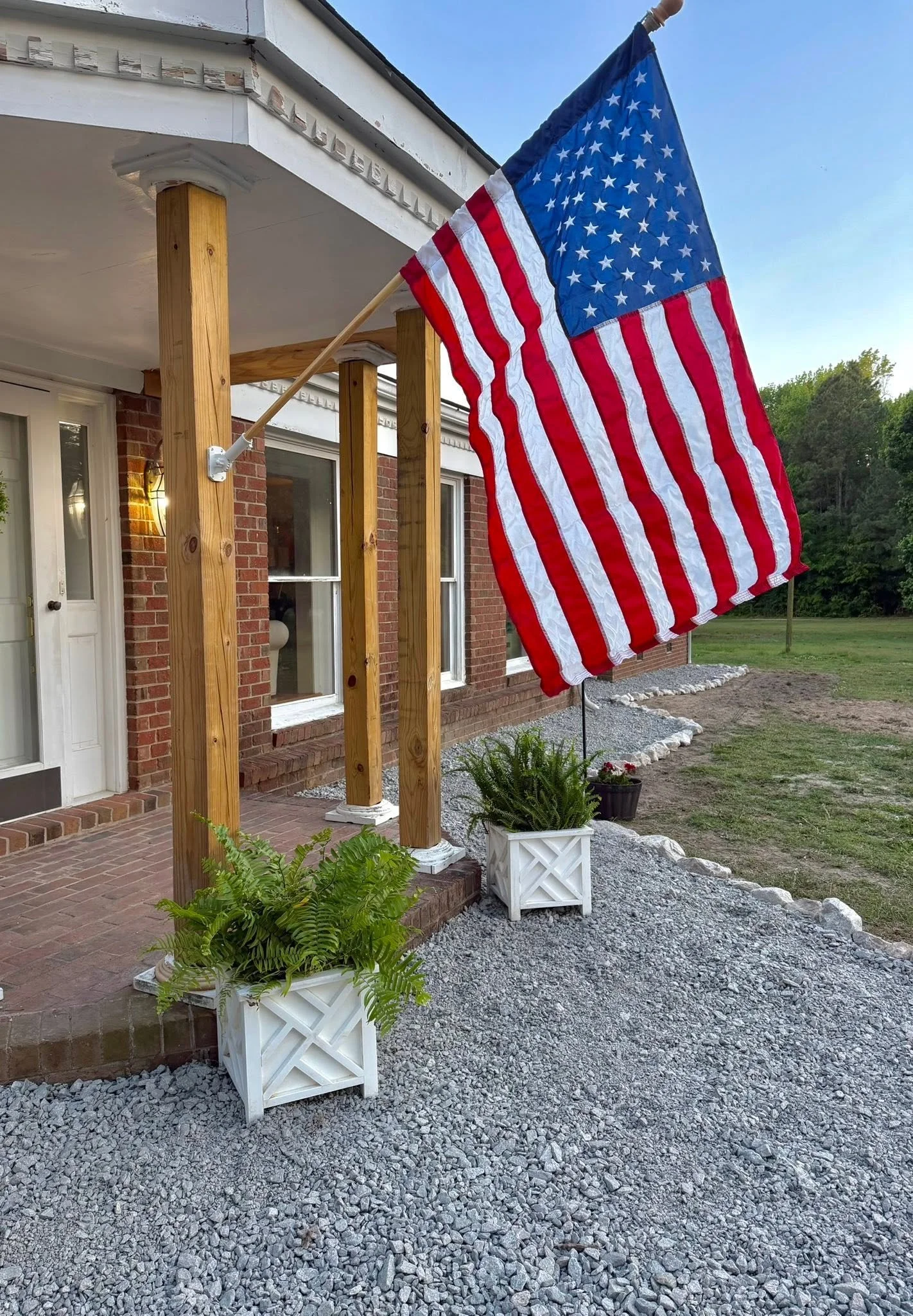American flag hanging outside a brick house with a white door and windows, decorated with new wooden posts in a sunny yard with gravel and plants.