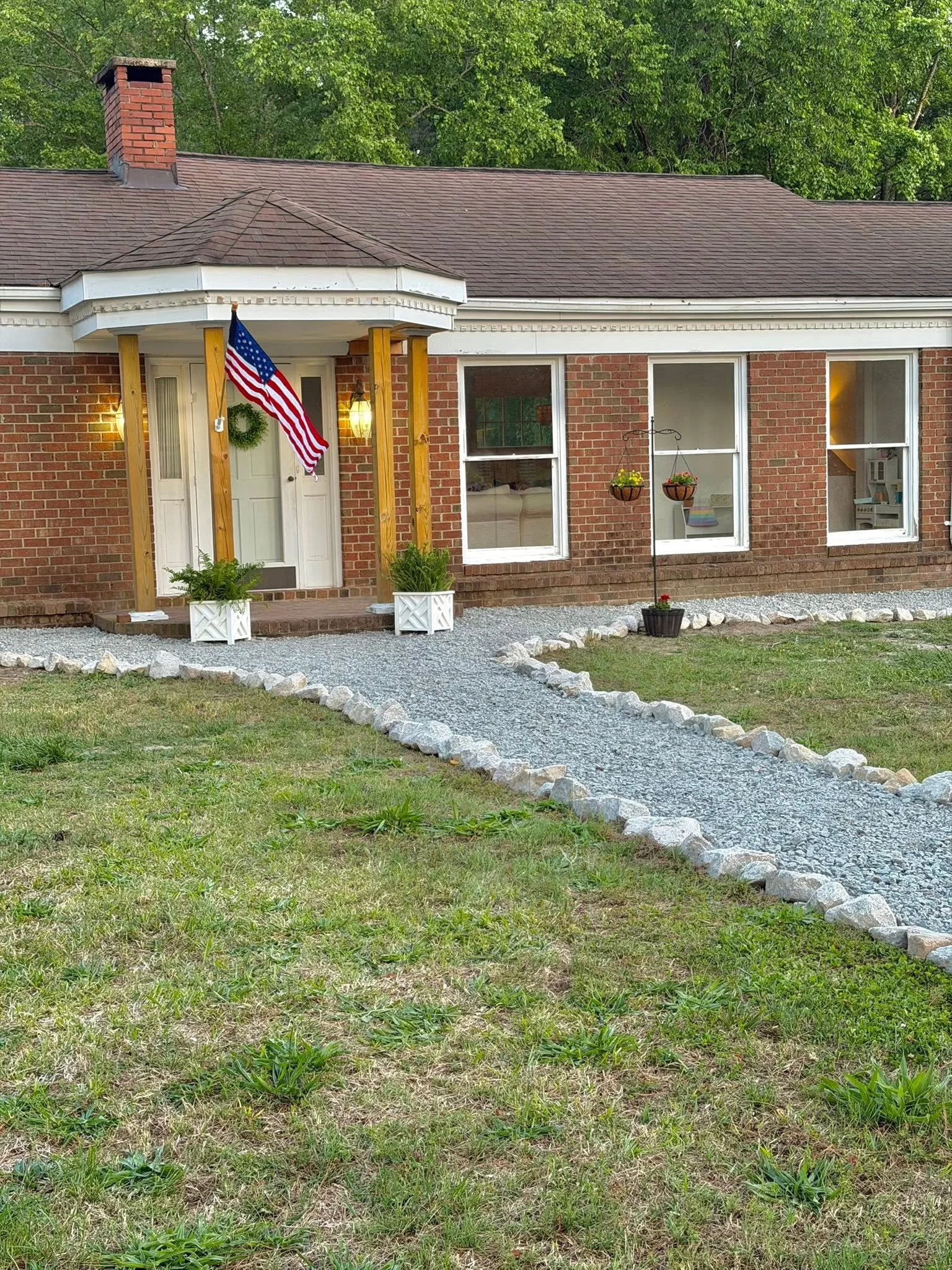A brick house with a front porch, American flag hanging near the door, and a curved gravel walkway leading to the entrance. The house has beige window frames, hanging flower baskets, and a green lawn with some rocks along the walkway. Trees with green leaves are visible in the background.