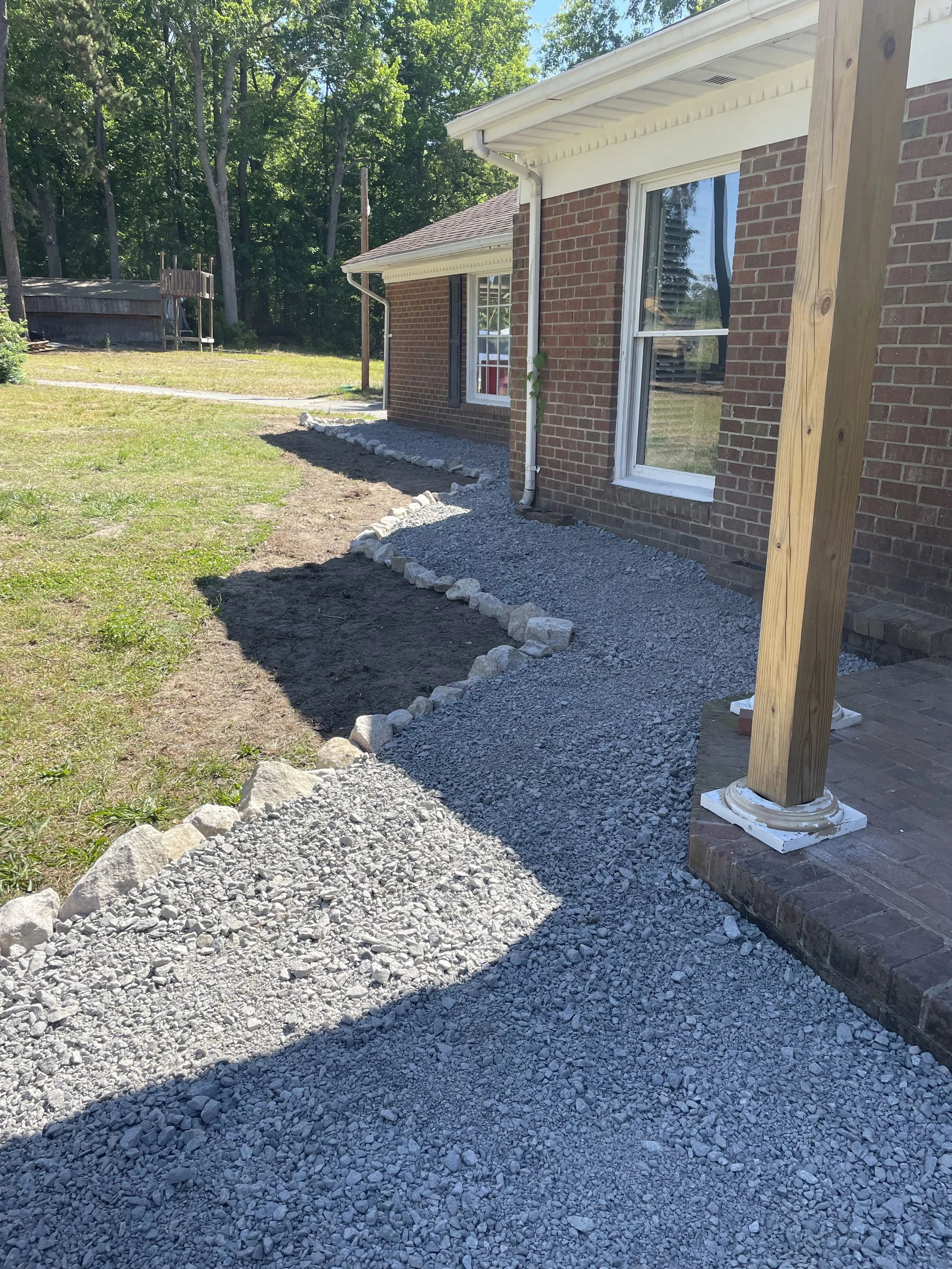 A backyard showing a brick house with a large window and a white gutter system, a new gravel pathway outlined with rocks, and a wooden post secured with a concrete base. The yard has patches of grass and is bordered by trees and a wooden fence in the background.