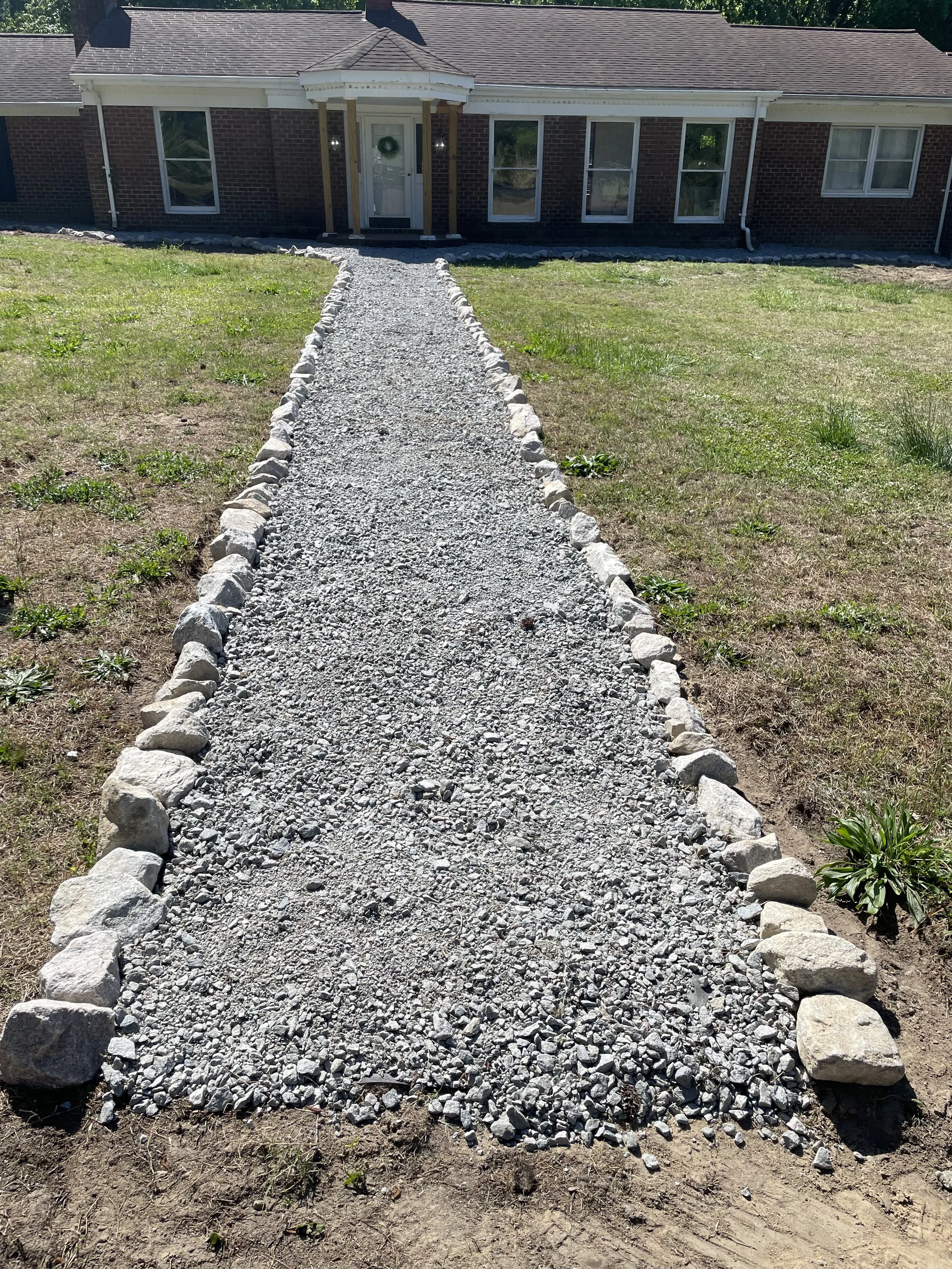 A gravel walkway lined with rocks leading to a house with a brick exterior, white trim, and a front porch with a wreath on the door.