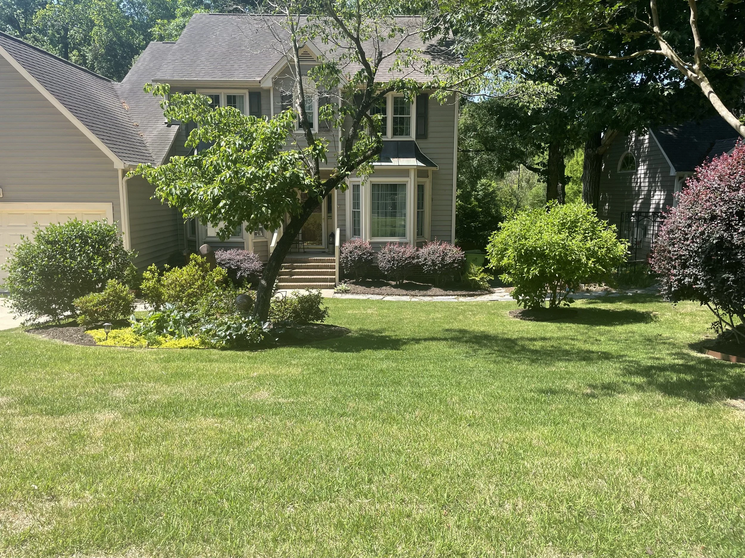 A two-story house with gray siding, surrounded by trees and bushes, with a well-maintained green lawn in the foreground.