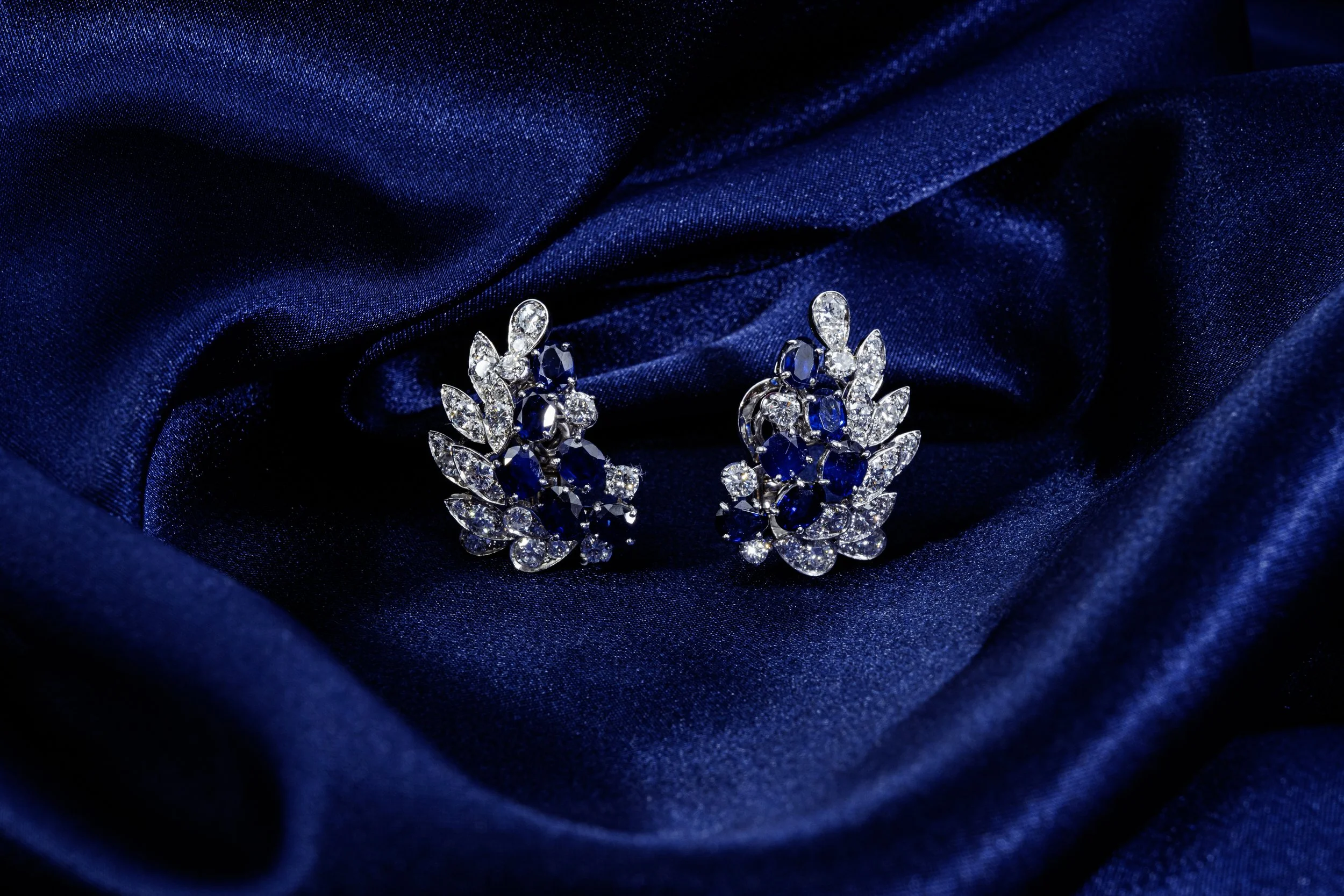 A pair of silver earrings with blue and clear gemstones, placed on dark blue satin fabric.