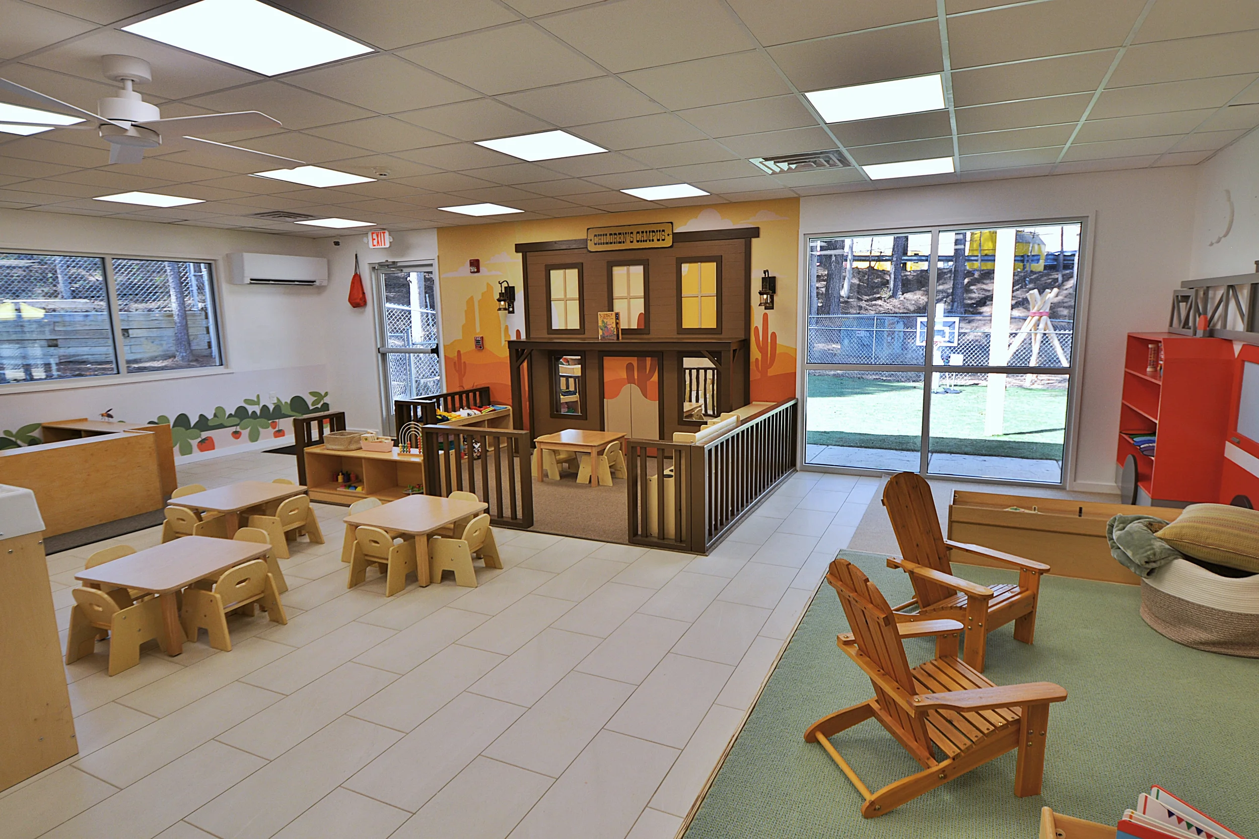 A spacious indoor play area with small tables and chairs, a playhouse labeled 'Children's Campus,' and outdoor playground equipment visible through large windows.