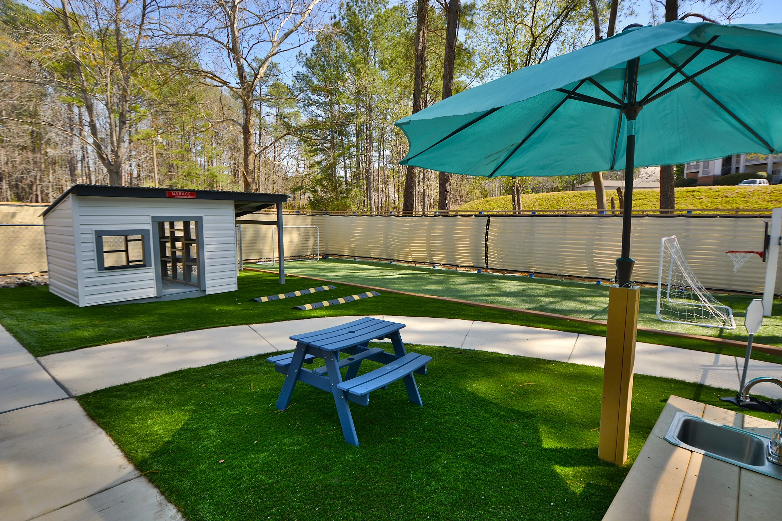 Backyard with artificial grass, picnic table, large blue patio umbrella, small white playhouse, soccer goal, and trees in background.