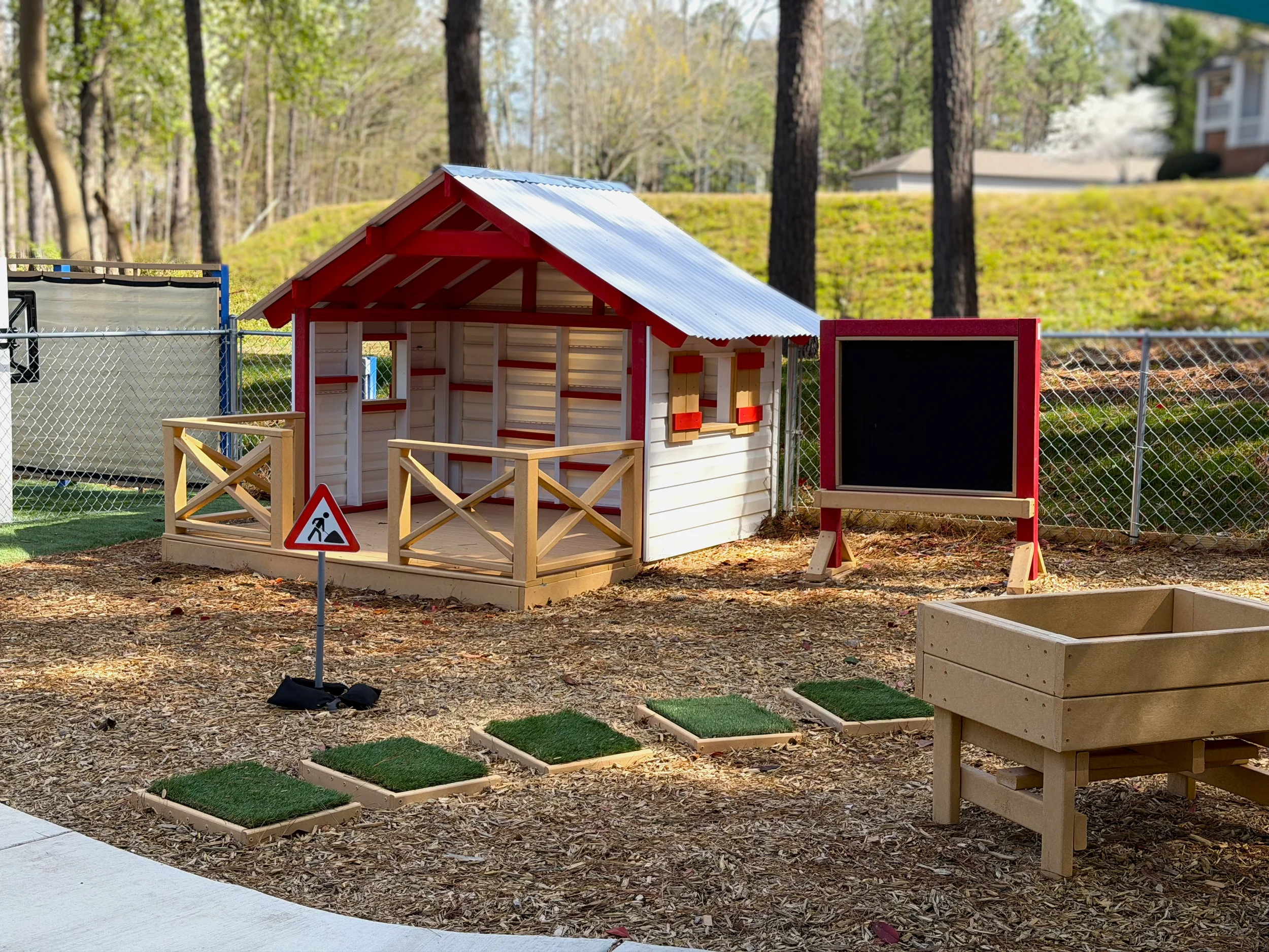 A small wooden outdoor playhouse with a corrugated metal roof, surrounded by a fence, located in a wooded park area with trees and houses in the background. There are signs and artificial grass mats in the play area.