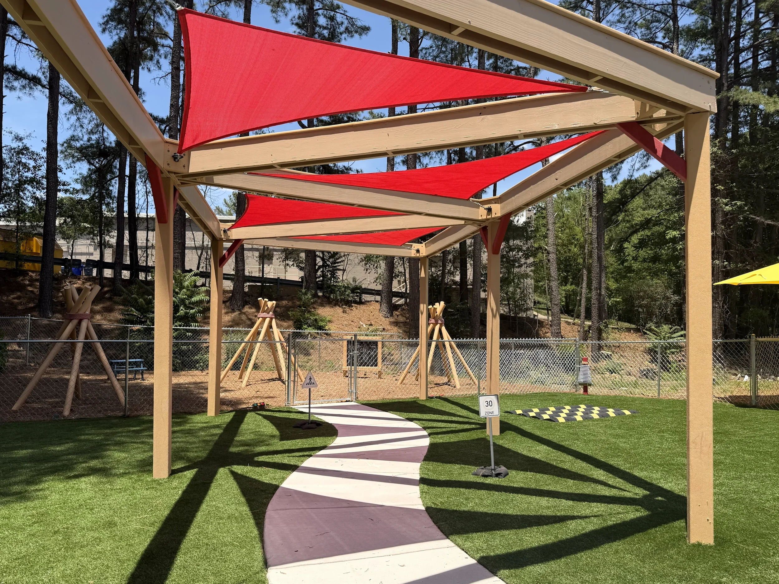 A playground with a wooden canopy structure featuring red fabric shades, a winding concrete pathway, a grassy area, and trees in the background.