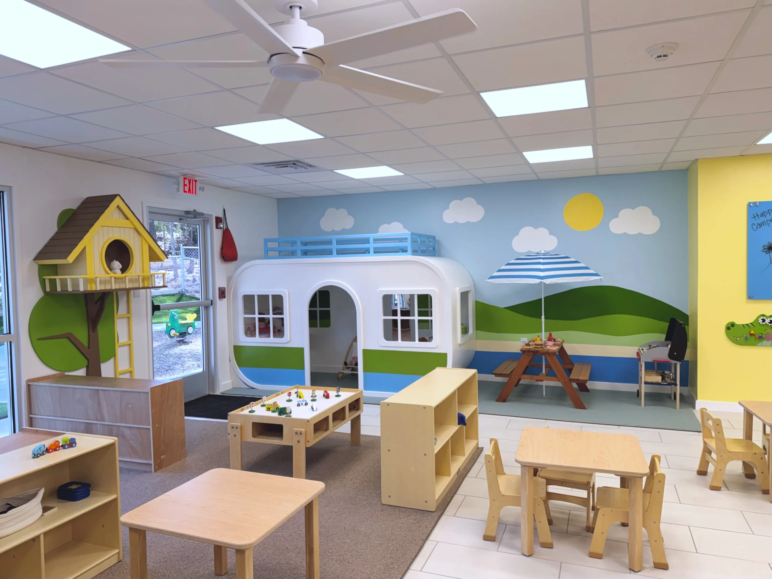 A colorful children's playroom with a painted mural of clouds, hills, and a sun on the wall. There are wooden tables and chairs, a small playhouse, toy vehicles, and a play table with building blocks. 