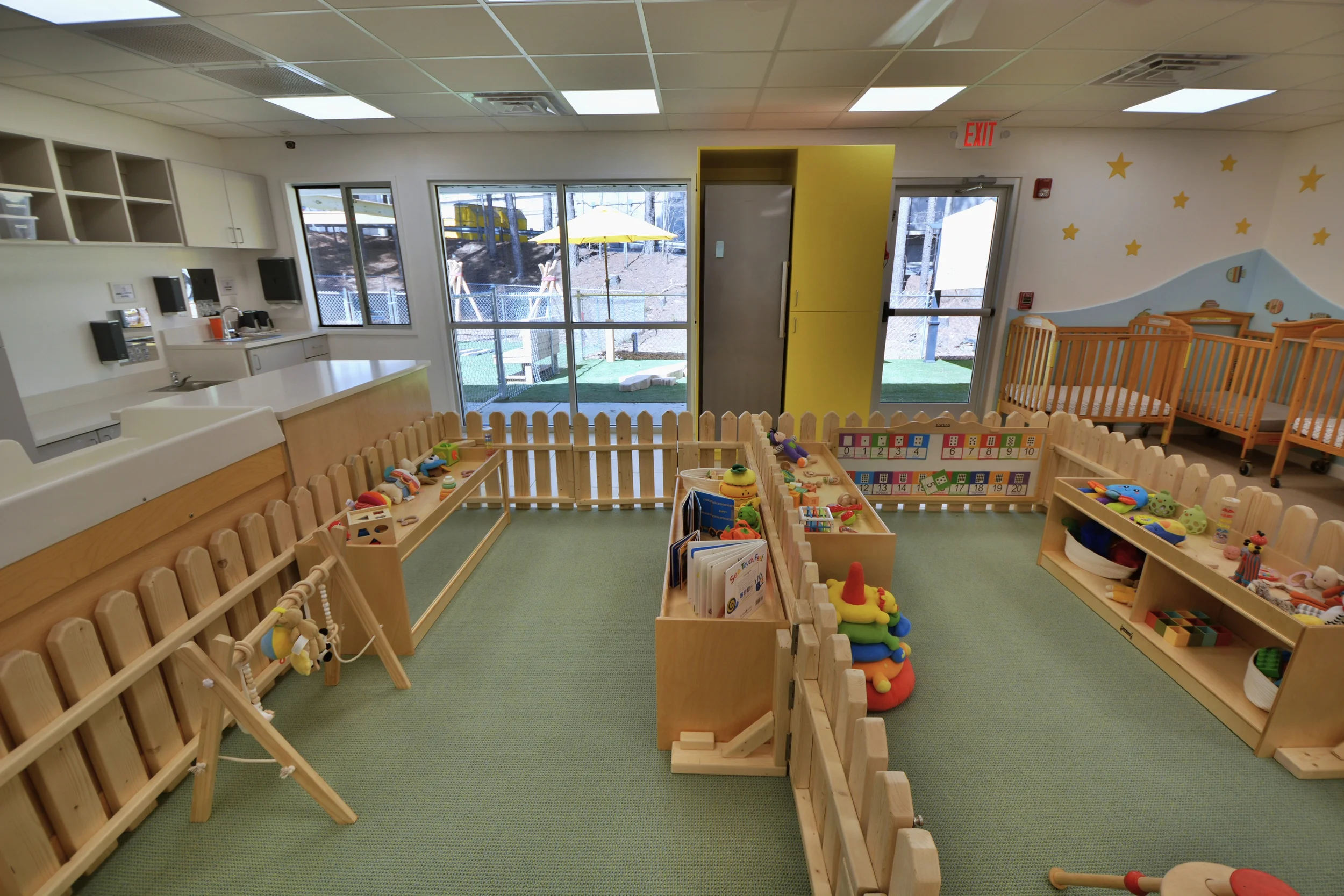 Interior view of a bright, colorful daycare or preschool classroom with wooden toys, books, and play areas. Large windows with a view of an outdoor playground with trees, and a fence. Walls decorated with stars and a mountain mural.